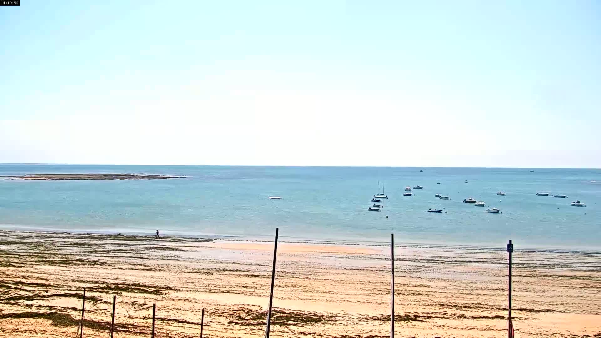 A sandy beach stretches to a calm, light-blue sea where several small boats are anchored under a clear sky.