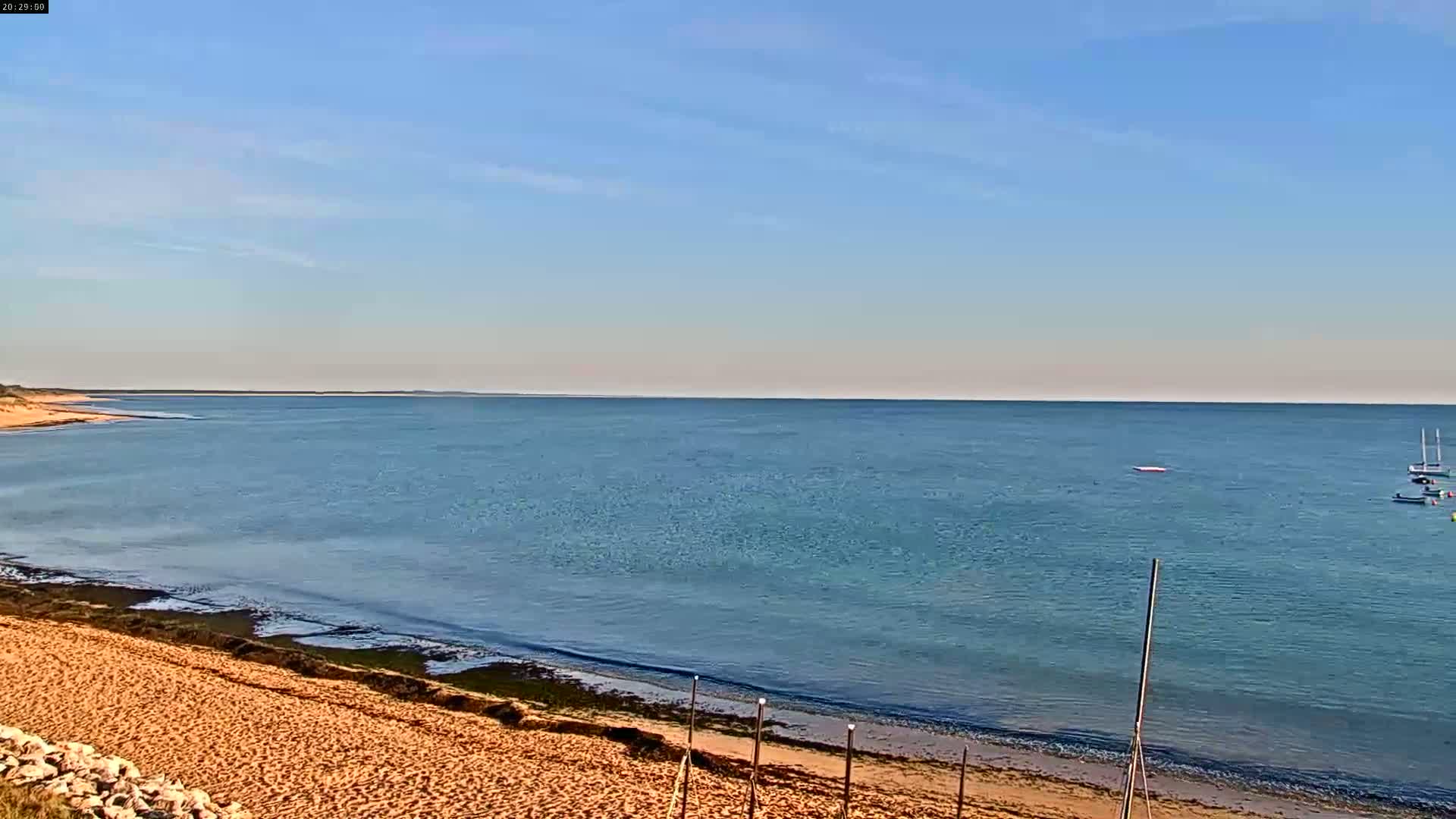A sandy beach slopes down to a calm, blue ocean under a clear, bright sky, with a few small boats visible in the distance.