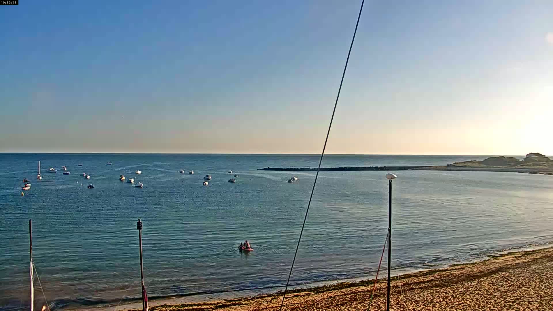 A calm, sunny day shows a sandy beach, several boats in the water, and a small landmass in the distance.