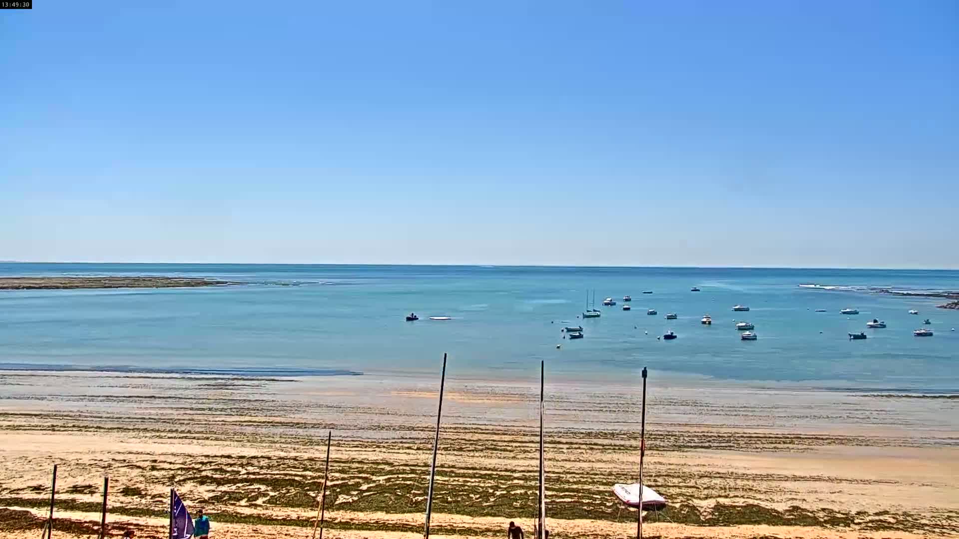 A sandy beach with several small boats in calm, shallow water under a clear blue sky.