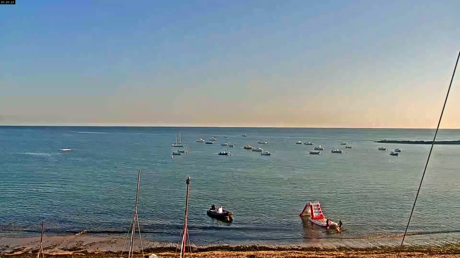 A calm, clear day reveals a sandy beach, several small boats on the water, and an inflatable water slide near the shore.