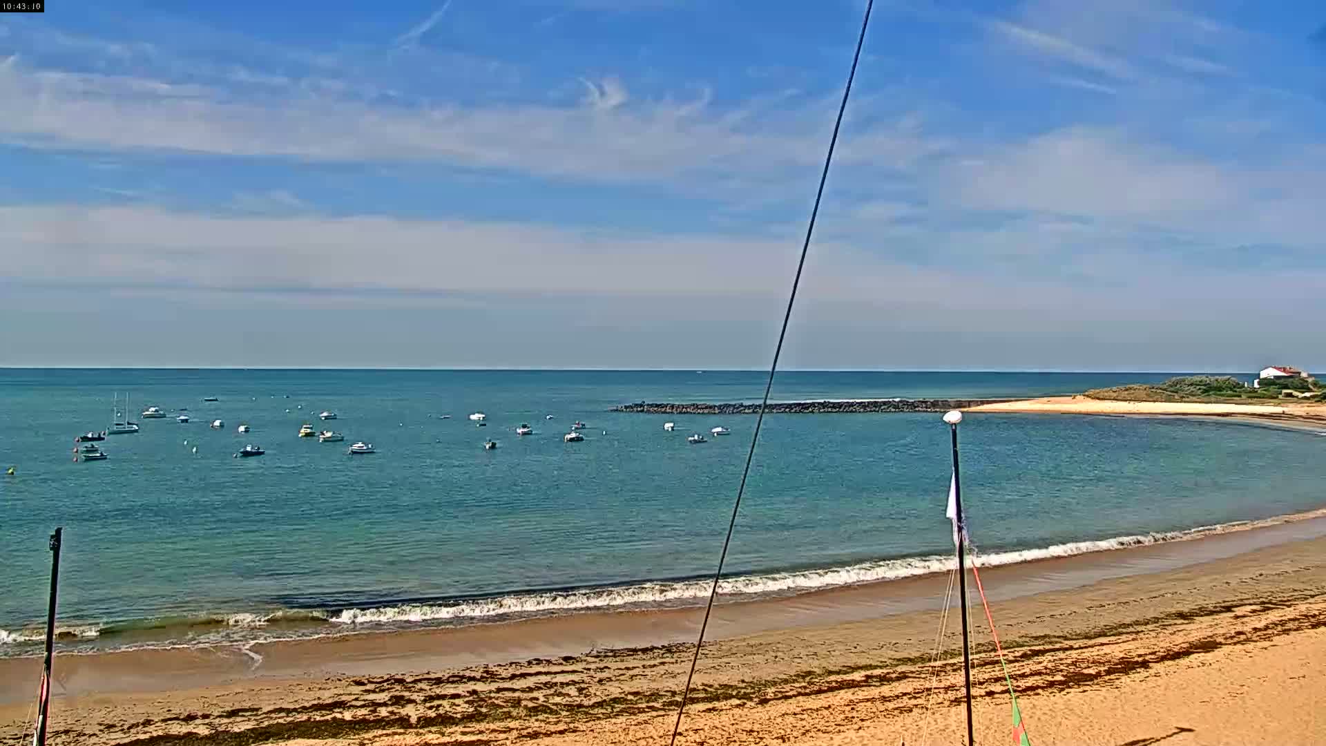 A sandy beach with gentle waves laps the shore, under a mostly sunny sky, with numerous small boats visible in the calm water beyond a short breakwater.