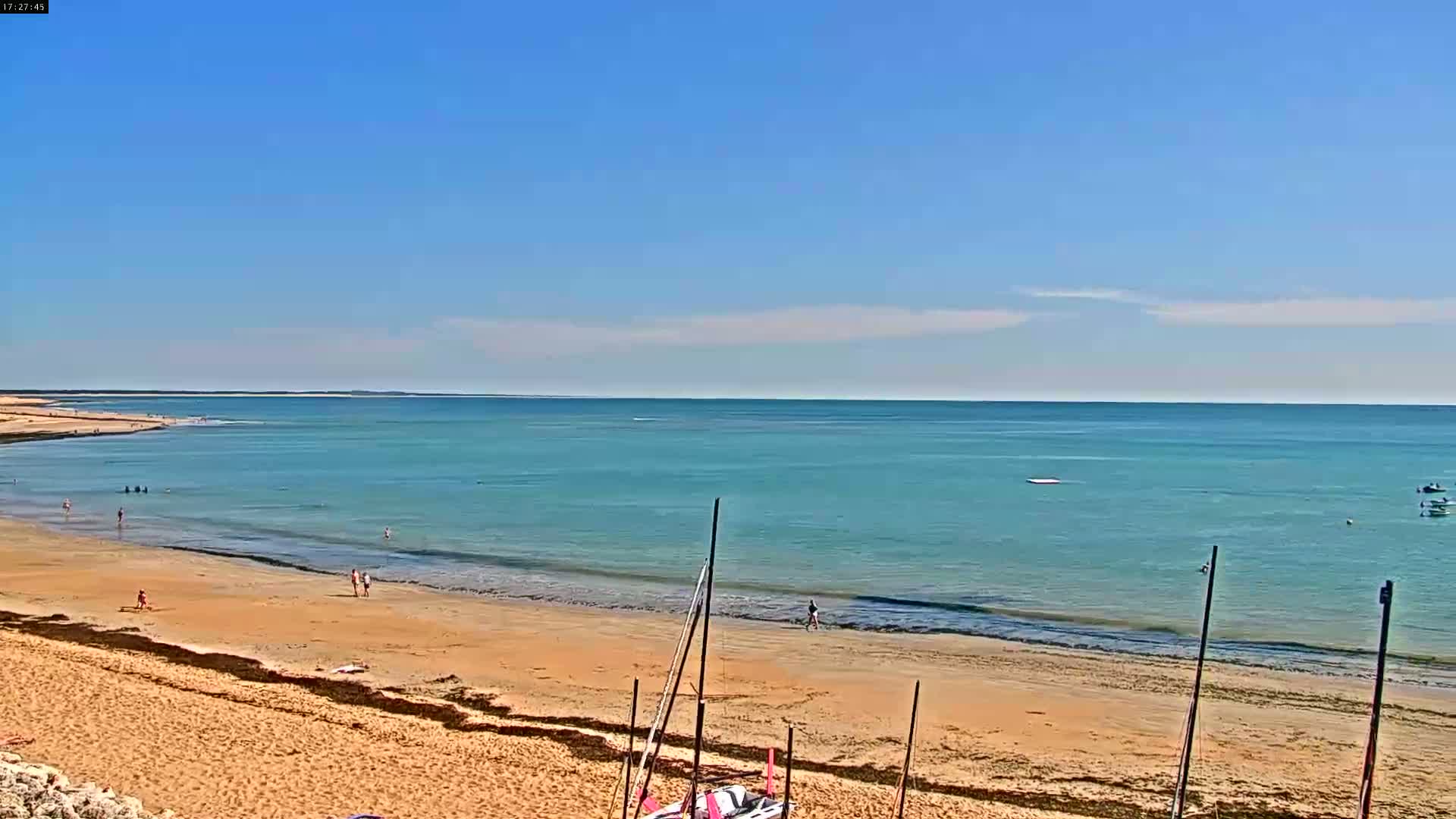 A sandy beach with a few people on it slopes down to a calm, light-blue ocean under a clear, sunny sky.