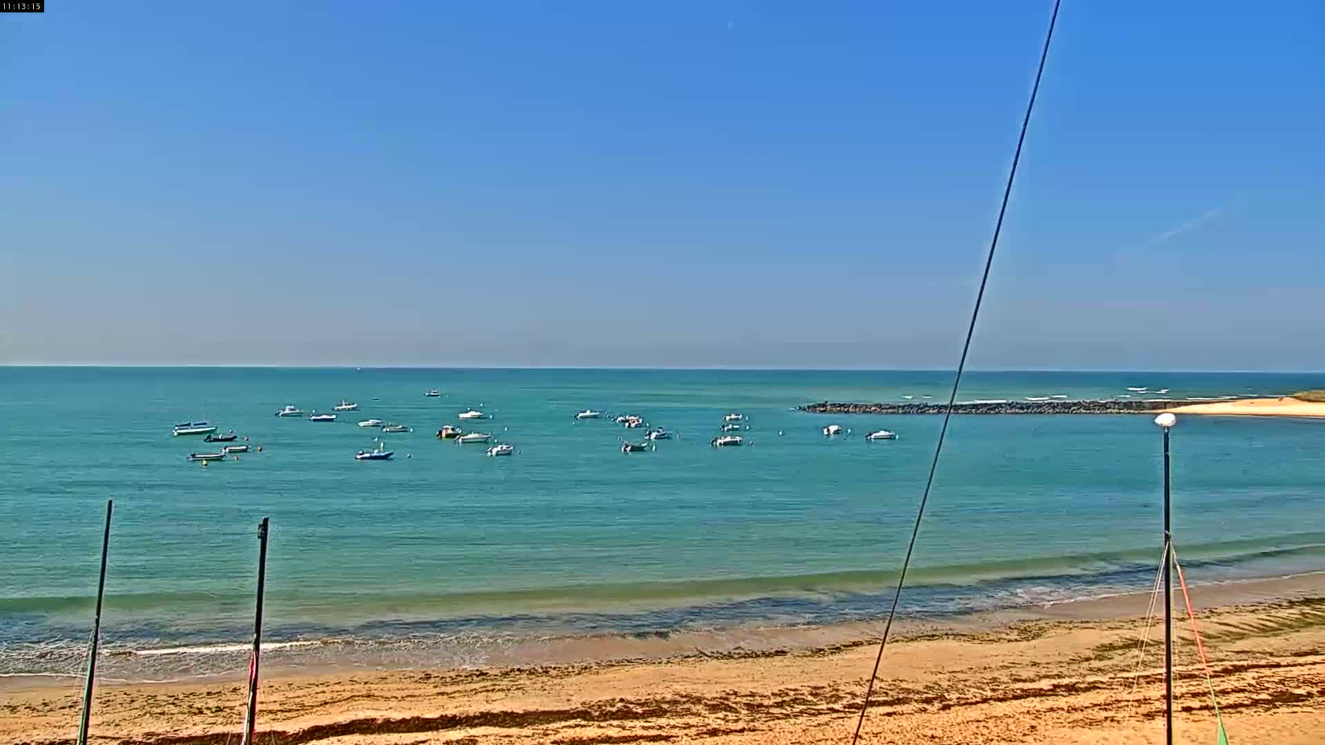 A sandy beach with calm, teal water and several small boats anchored in a bay under a clear blue sky.