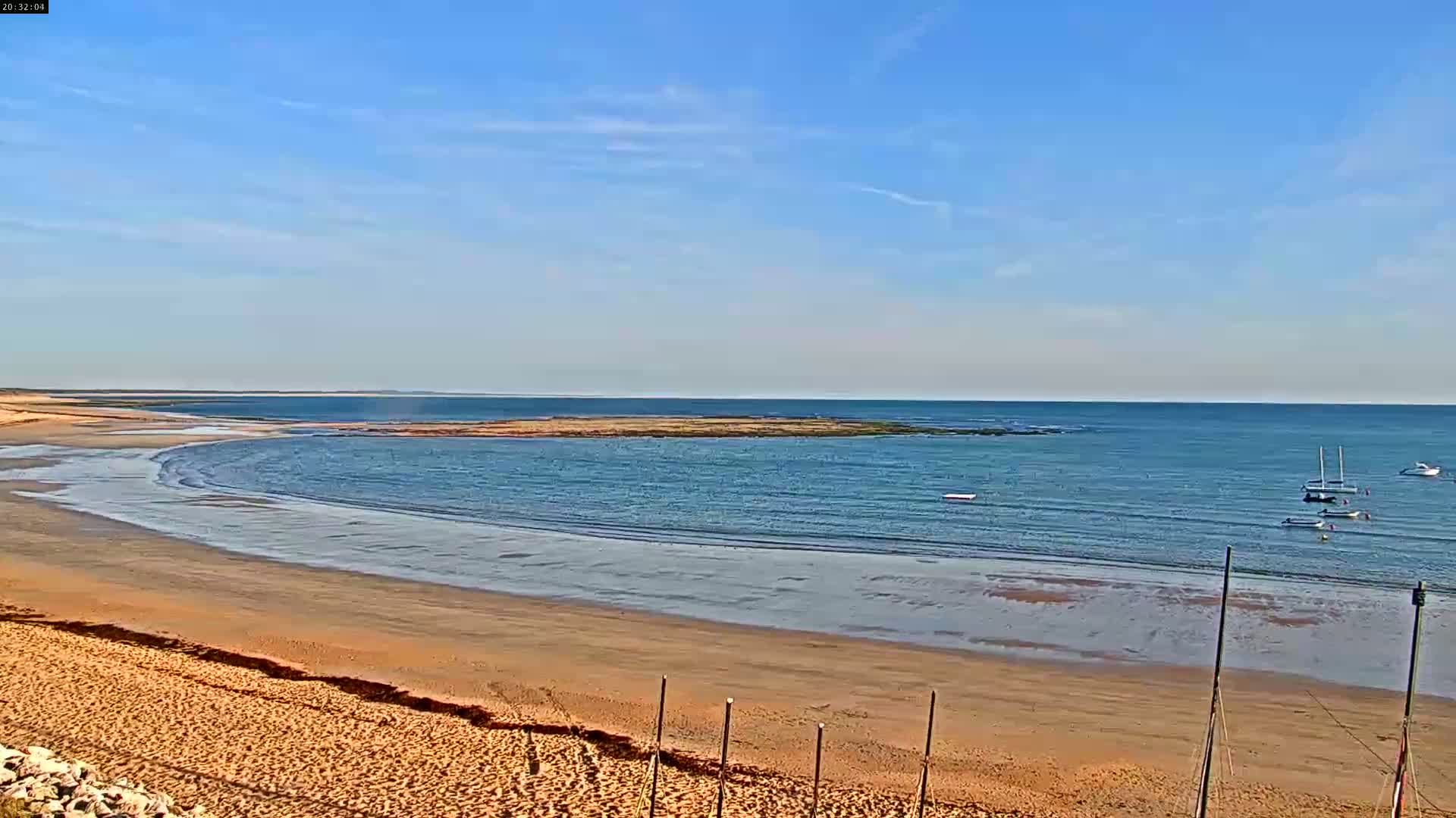 A sandy beach curves around a calm bay under a clear blue sky, with several small boats visible in the distance.