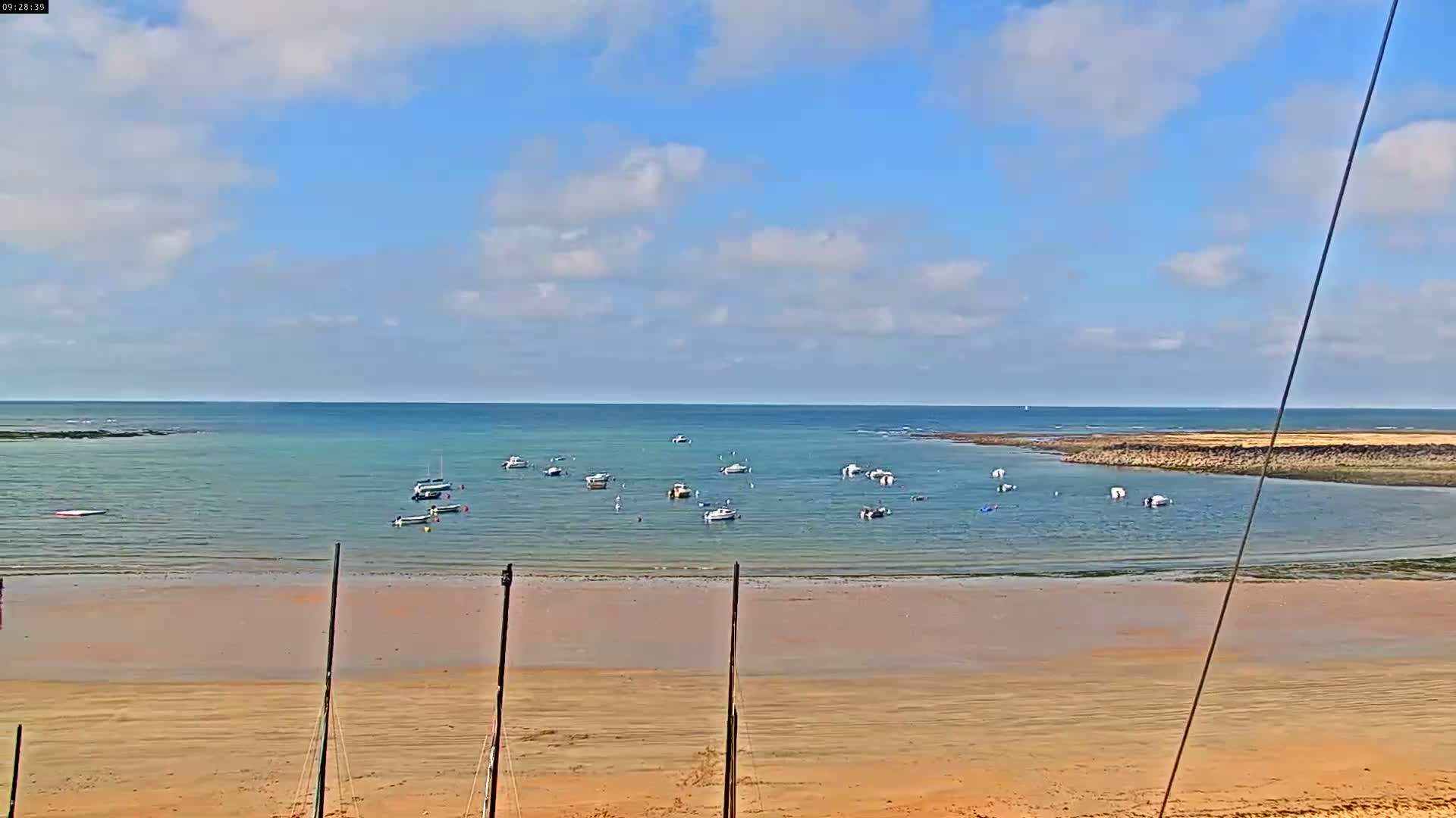 A sandy beach stretches out to a calm bay dotted with small boats under a partly cloudy sky.