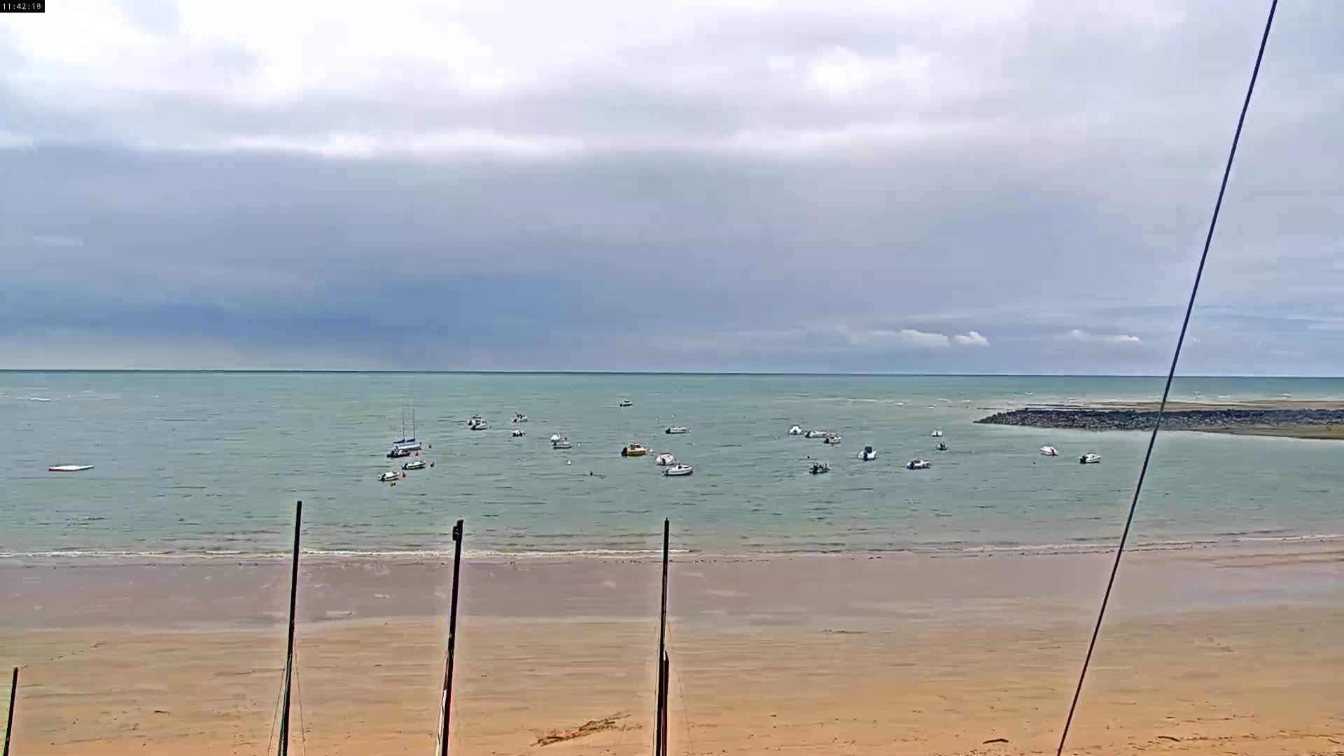 Numerous small boats are anchored in calm, shallow water near a sandy beach under a mostly cloudy sky.