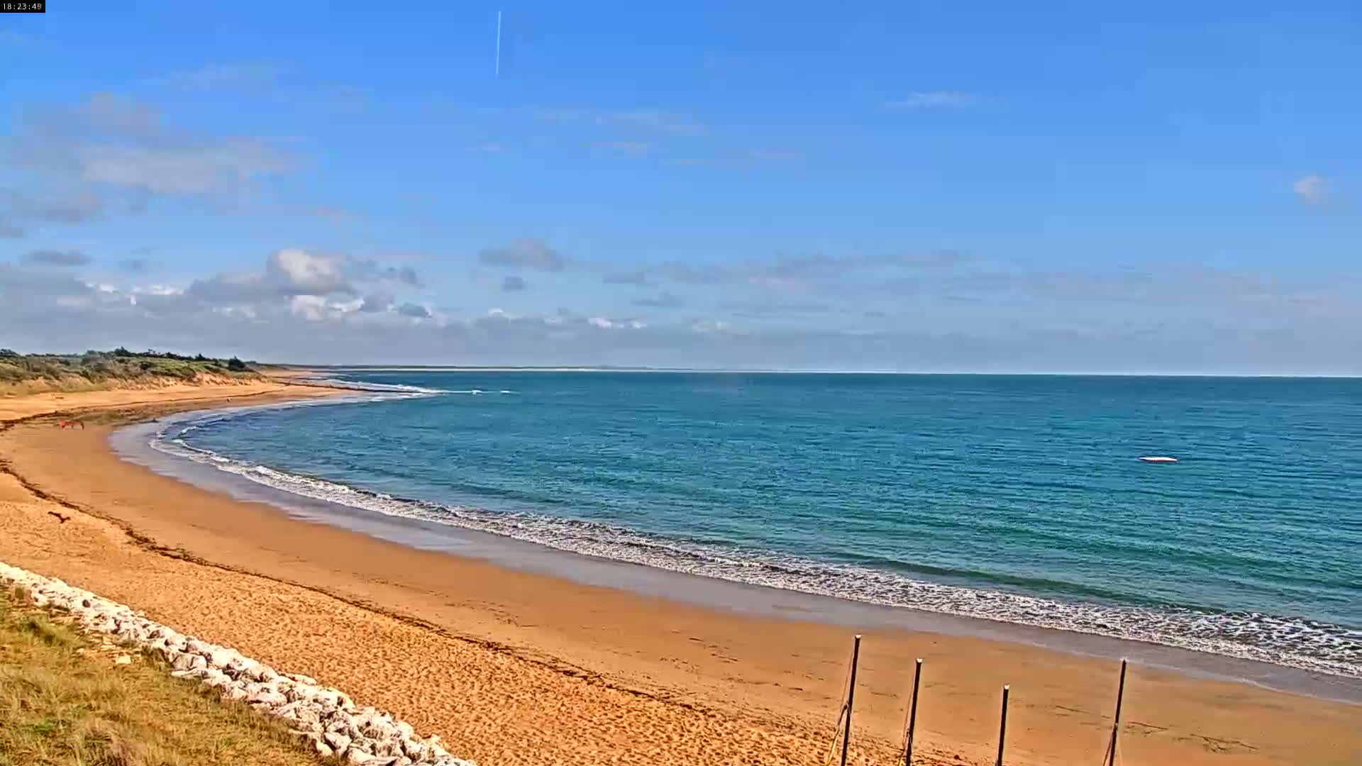 A sandy beach curves along a calm, teal ocean under a mostly sunny sky with scattered clouds.
