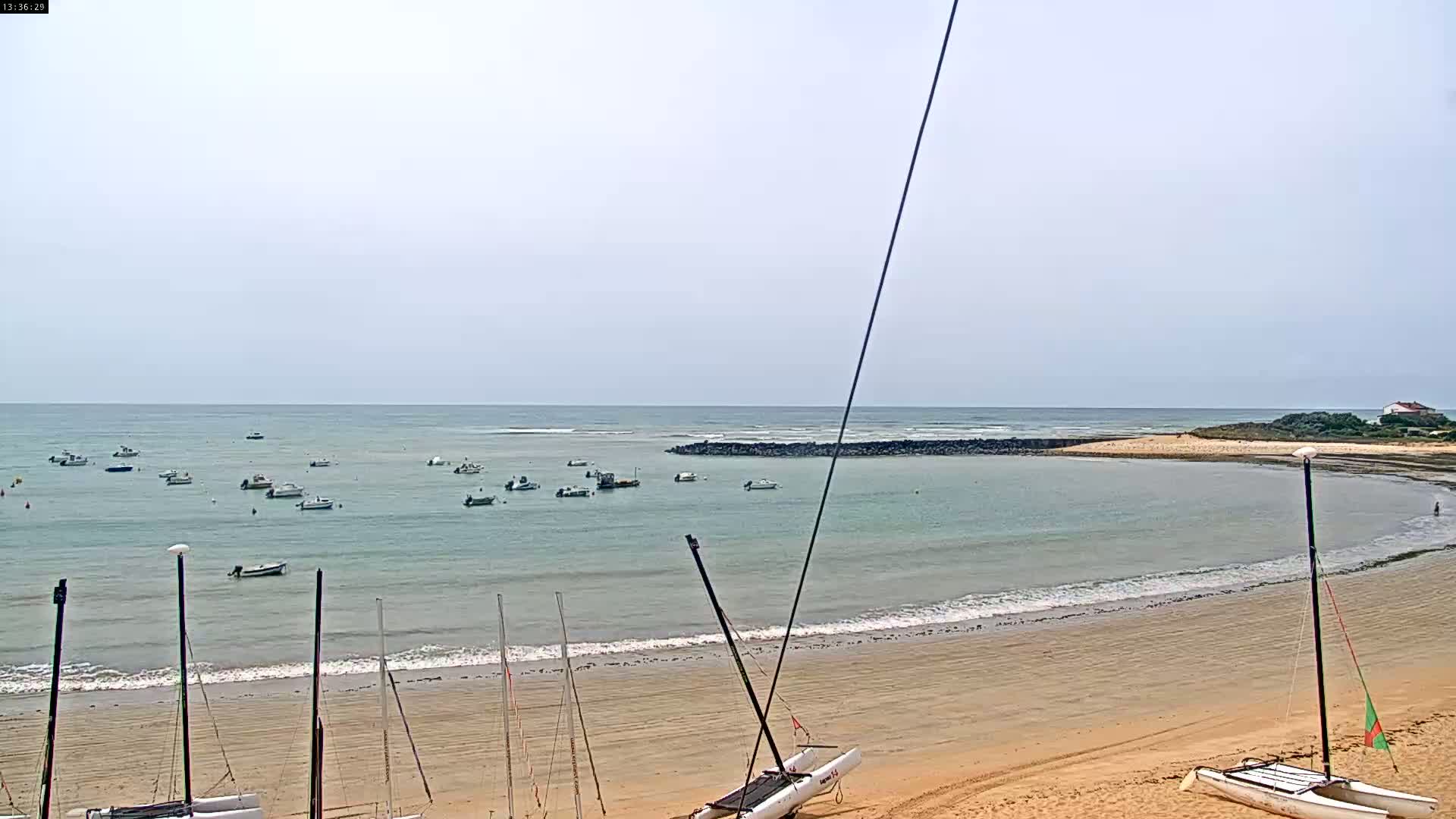 A sandy beach with several small sailboats on the shore and numerous small motorboats in calm, light-grey water under a mostly clear sky.