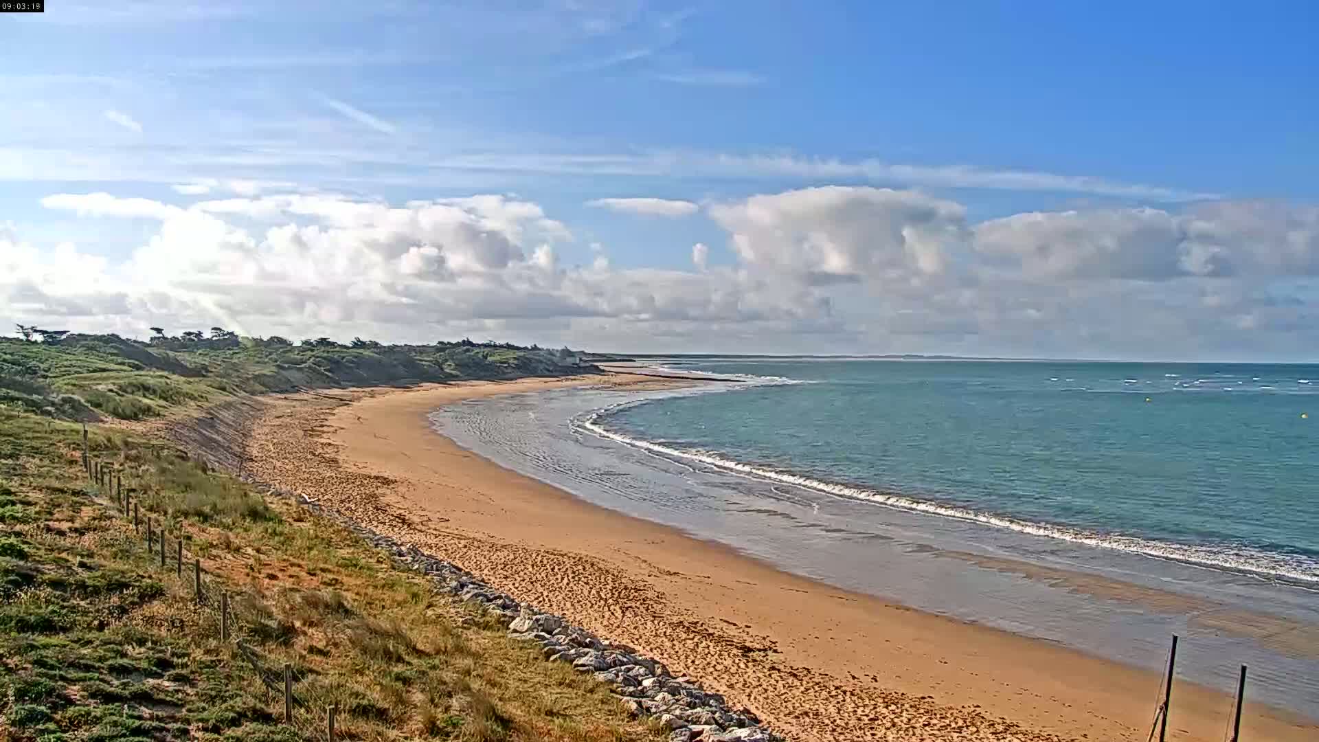 A sandy beach curves gently around a calm, blue ocean under a partly cloudy sky.