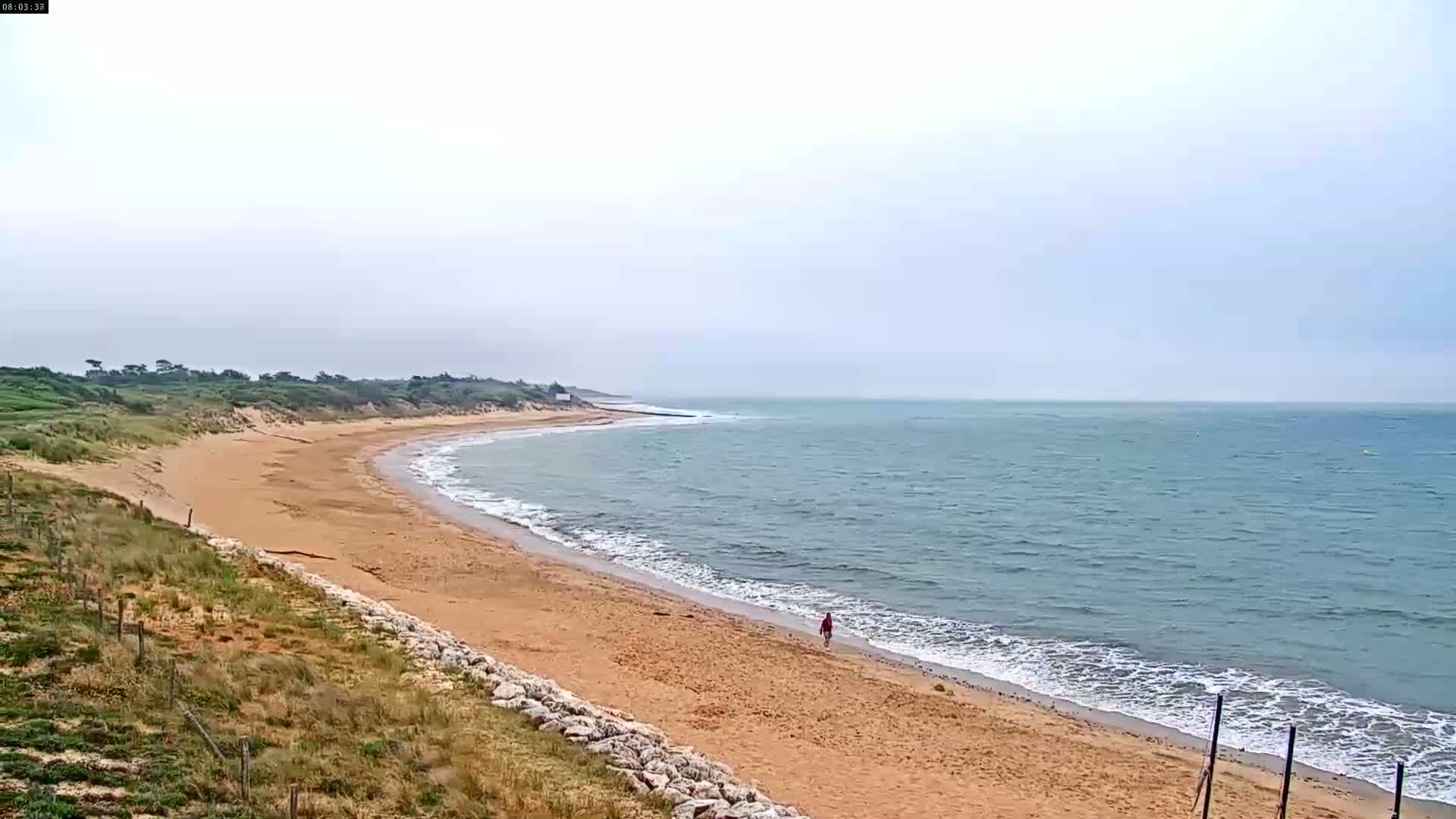 A person walks along a sandy beach that curves around a calm, light-blue ocean on an overcast day.