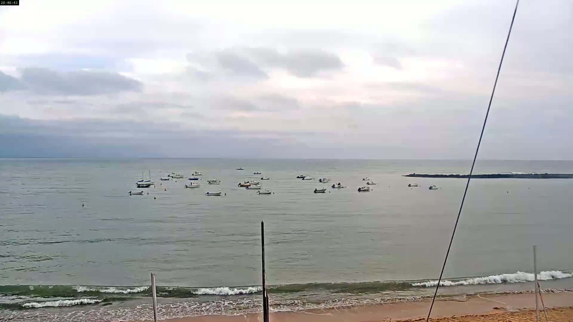 Many small boats are anchored in calm, grey water under a cloudy sky near a sandy beach with small waves.