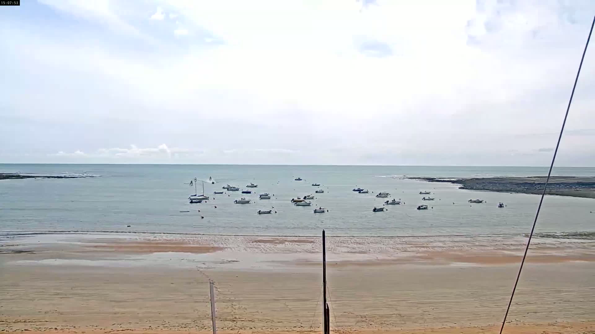 A sandy beach stretches to a calm body of water where numerous small boats are anchored under a mostly cloudy sky.