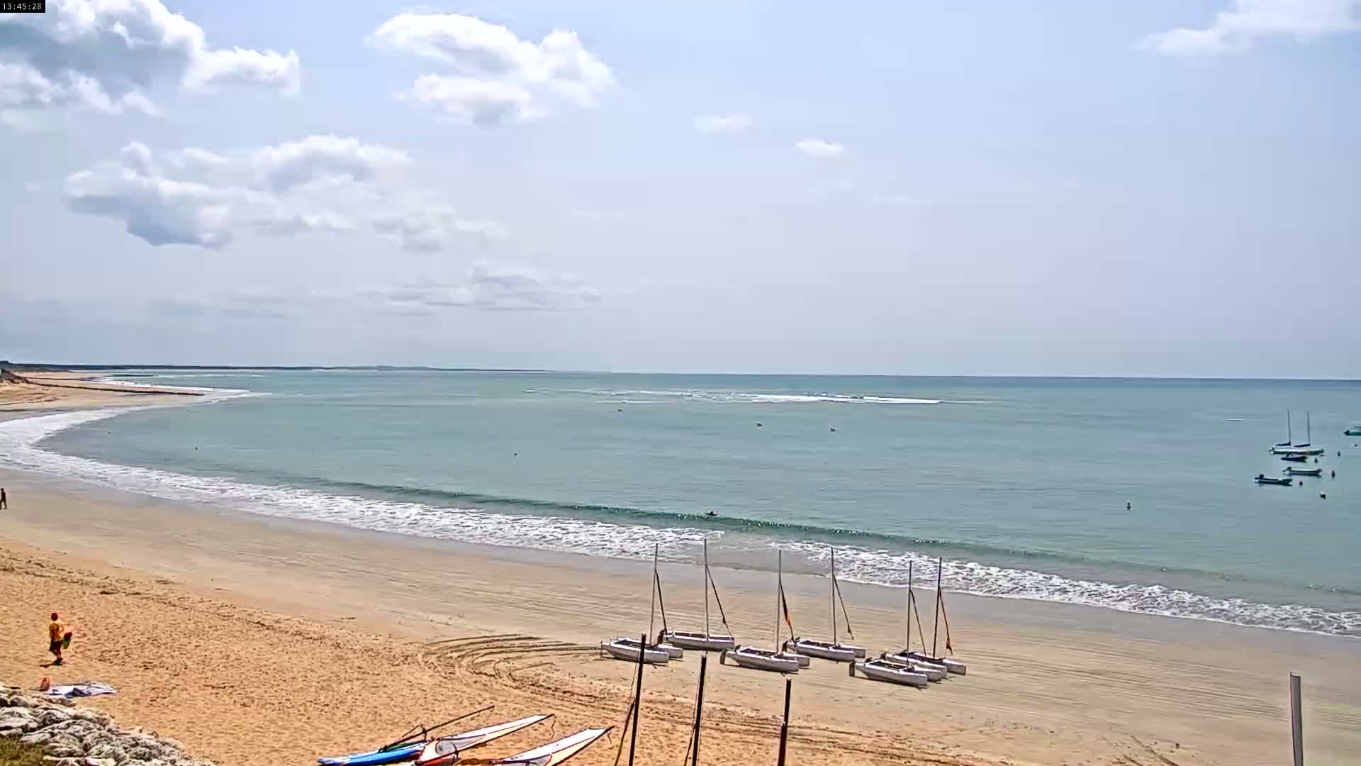 A sandy beach with small waves under a partly cloudy sky has several sailboats on the shore and a few more in the calm water.