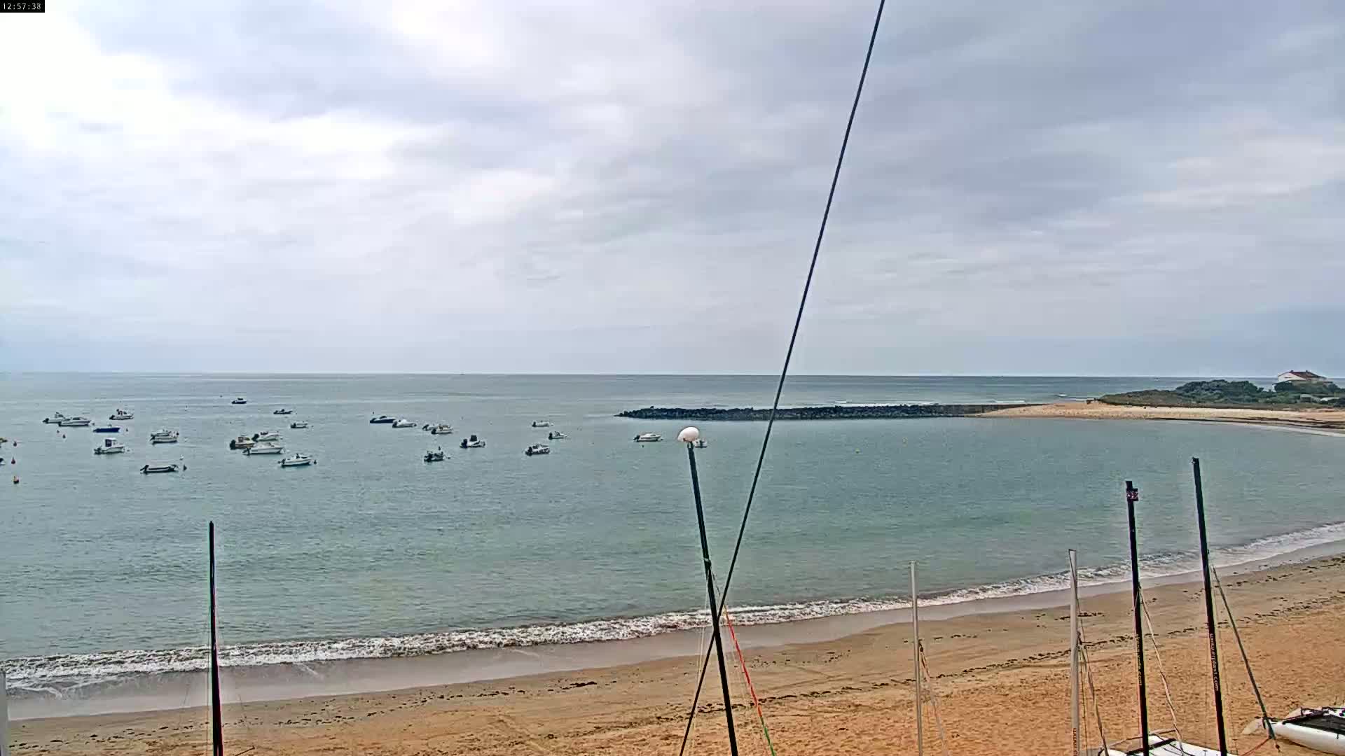 A sandy beach with several sailboats on the shore and numerous small boats in calm, overcast waters.