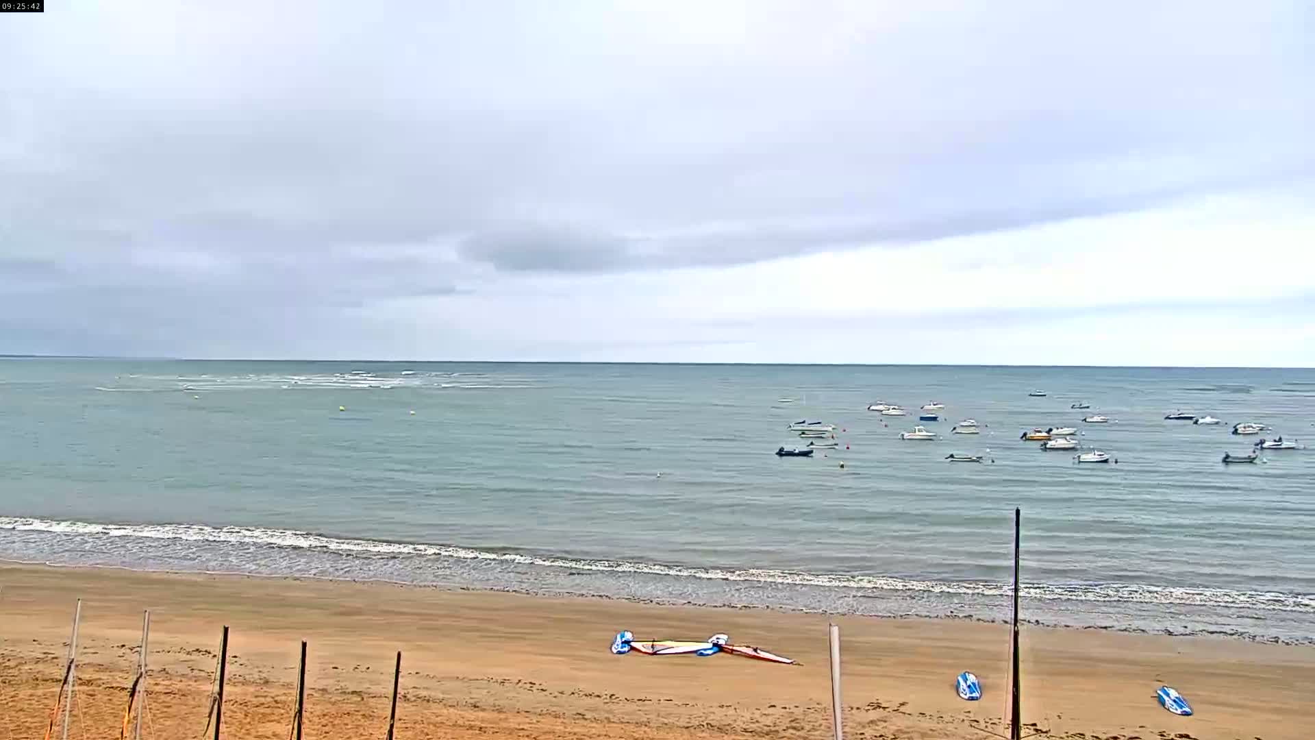 A sandy beach with several sailboards on the sand overlooks a calm sea with numerous small boats under an overcast sky.
