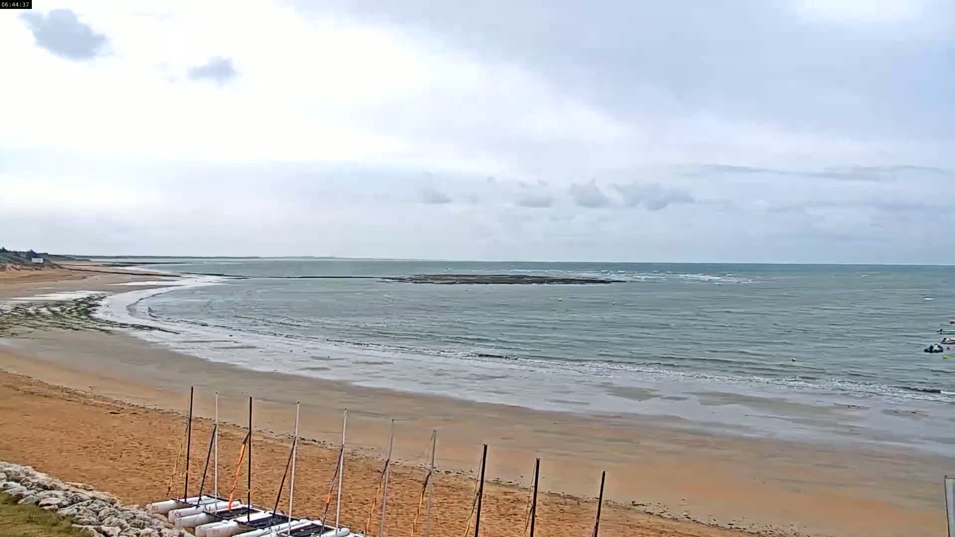 A sandy beach with several small sailboats on the shore stretches toward a calm, gray sea under a cloudy sky.