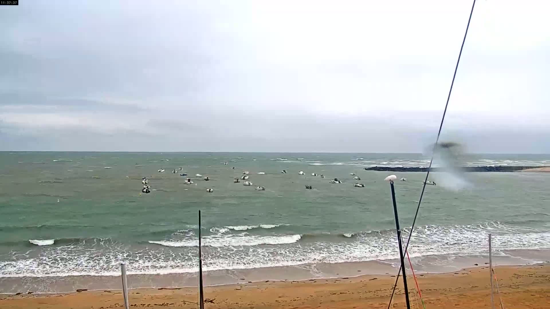 Numerous small boats are anchored in a calm, green-grey sea under an overcast sky, with small waves lapping a sandy beach.