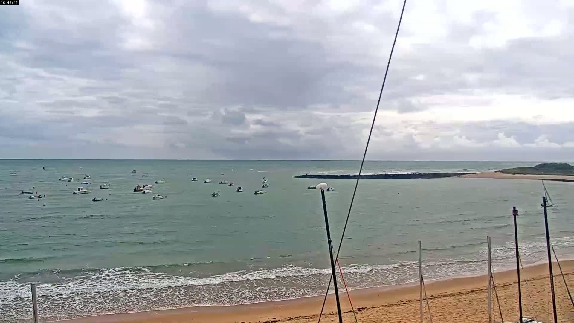 Numerous small boats are anchored in calm, greenish-grey water near a sandy beach under an overcast sky.