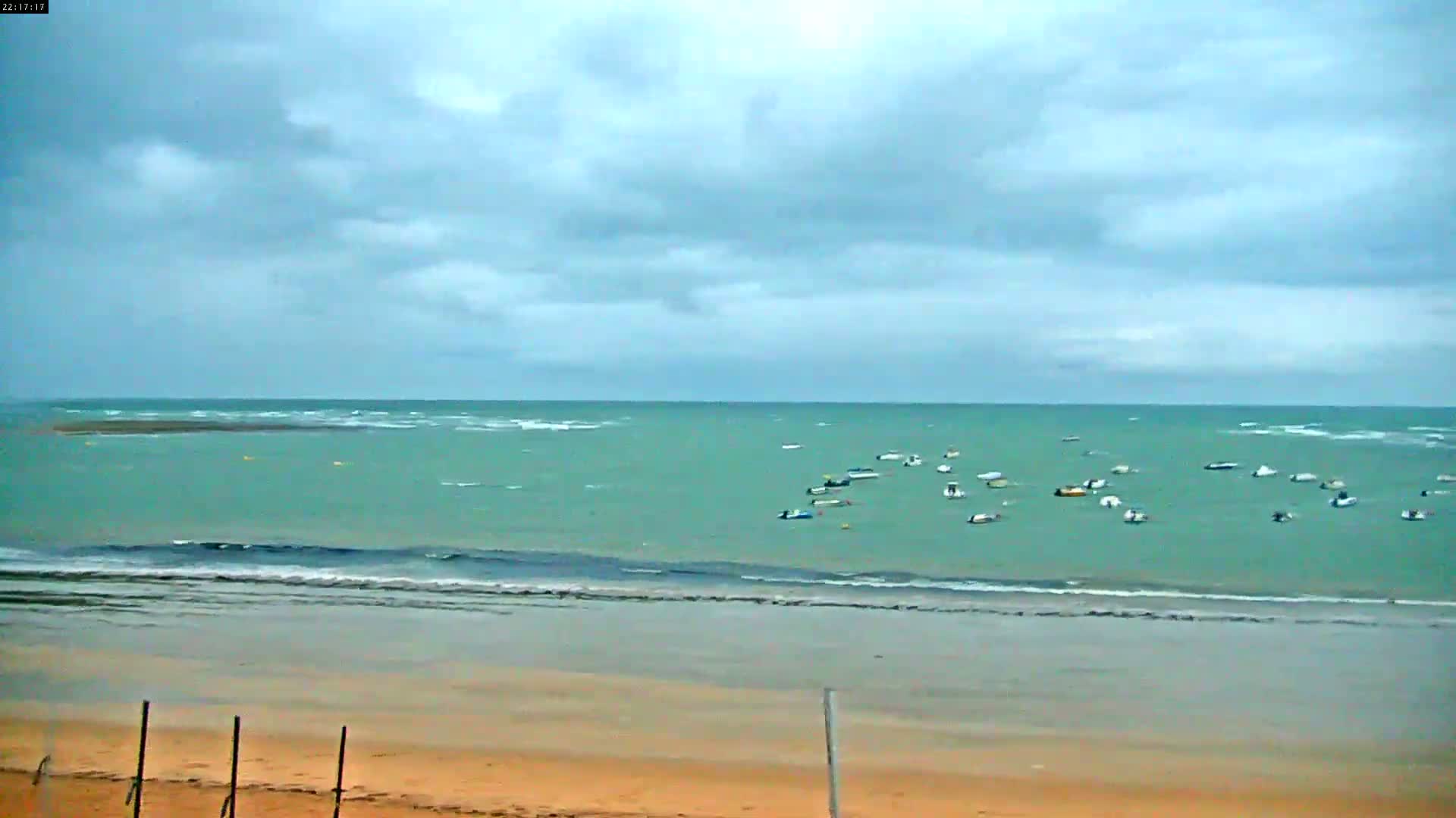 A sandy beach with a calm sea dotted with numerous small boats under a mostly cloudy sky.