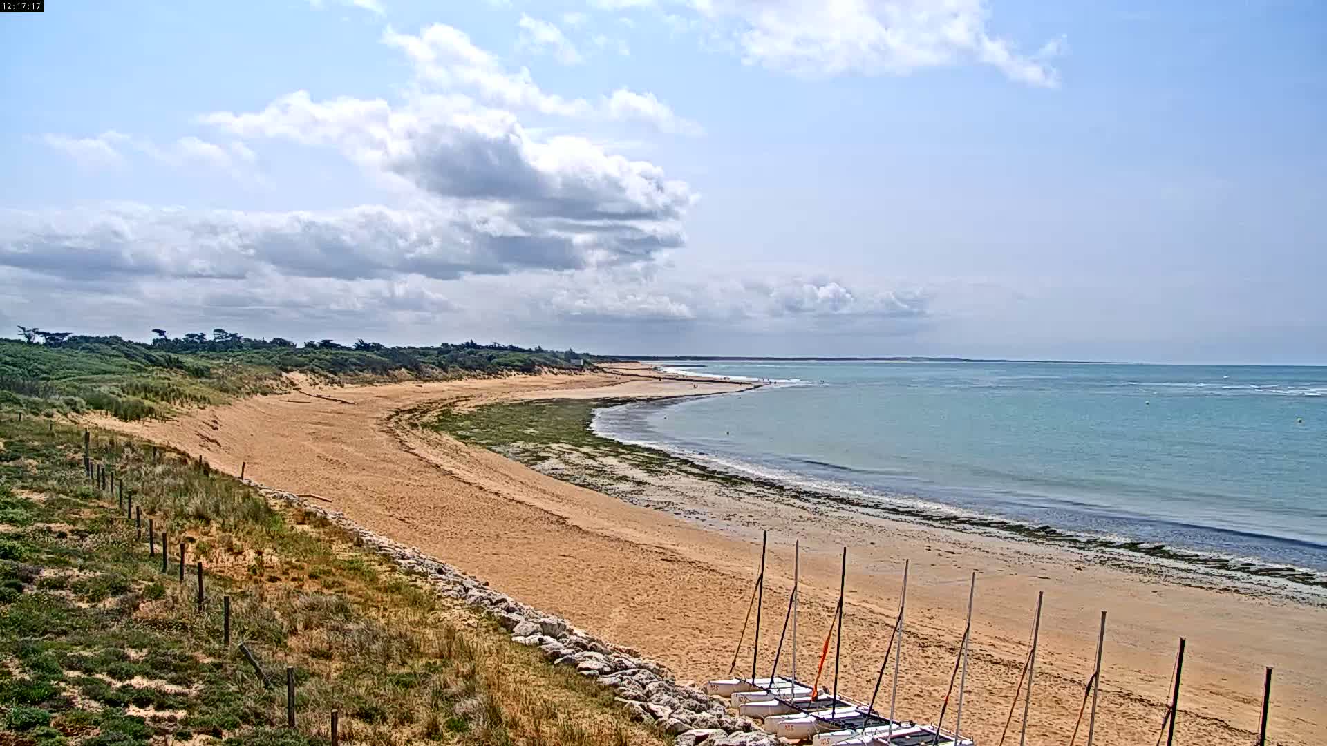 A sandy beach curves gently to meet a calm, light-blue ocean under a partly cloudy sky.