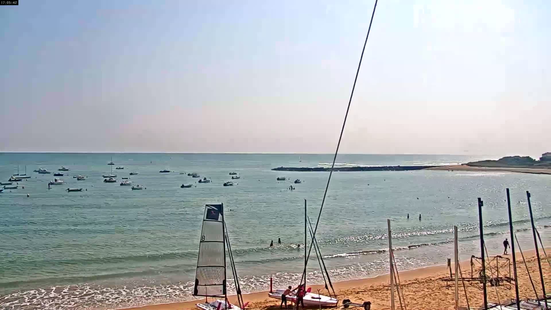 A sandy beach with several sailboats on the shore and numerous small boats in calm, clear water under a sunny sky.