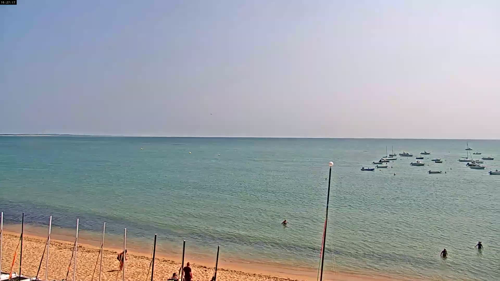 A sandy beach with several people and boats in calm, clear water under a bright, mostly clear sky.
