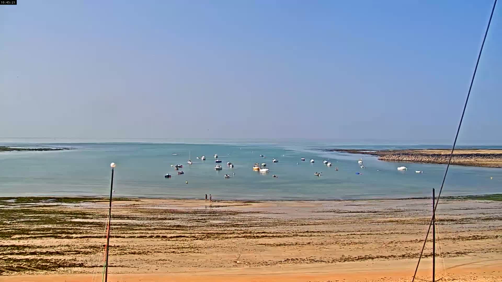 A sandy beach slopes down to a calm, light-blue bay where several small boats are moored under a clear, bright sky.
