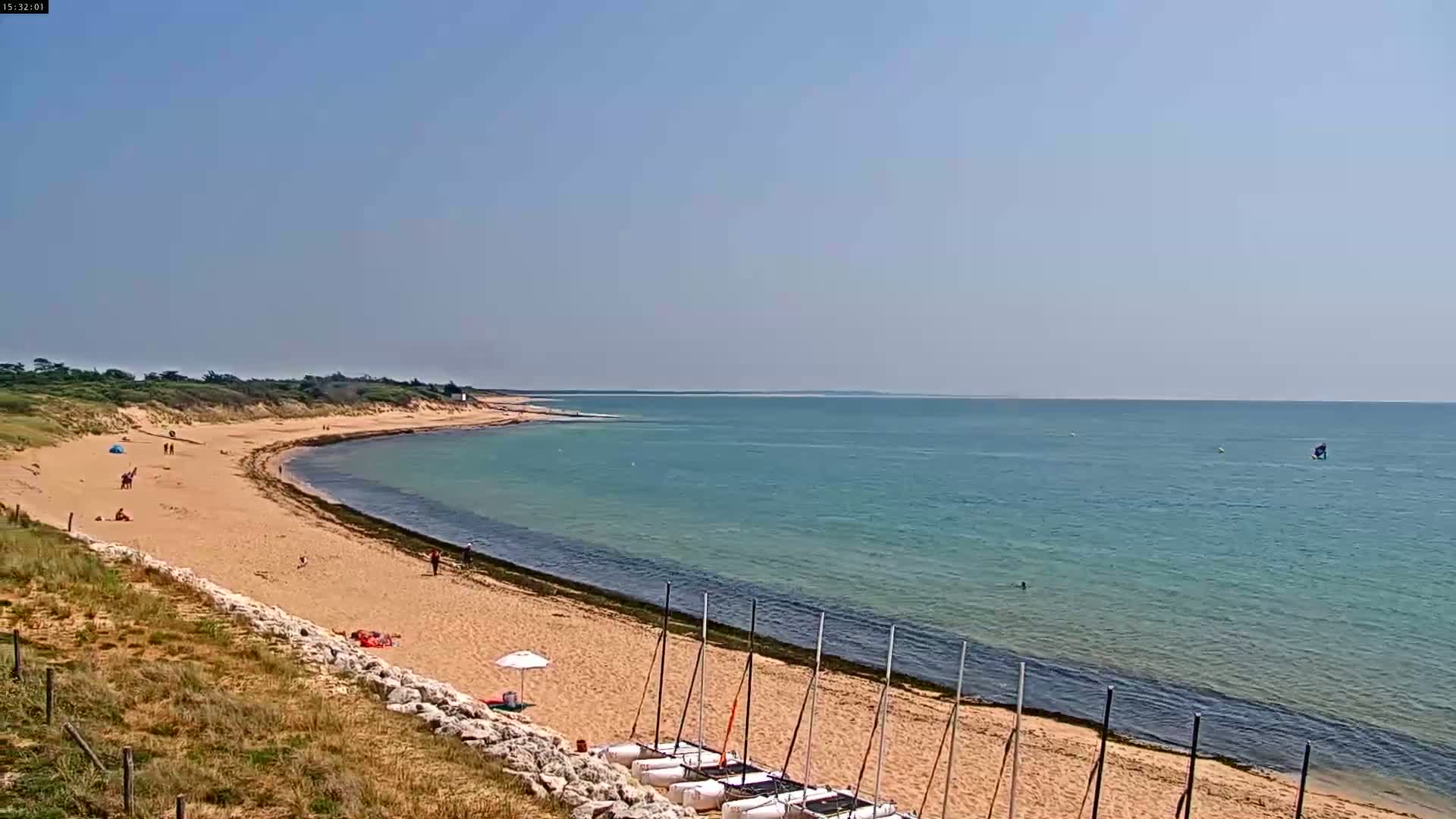 A sandy beach curves gently into a calm, teal ocean under a clear blue sky, with a few people scattered along the shore and several small sailboats lined up on the sand.