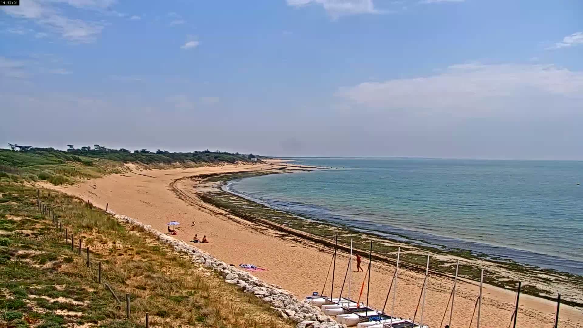 A sandy beach curves along a calm, teal ocean under a mostly sunny sky, with several small sailboats beached at the bottom and a few people relaxing on the sand.