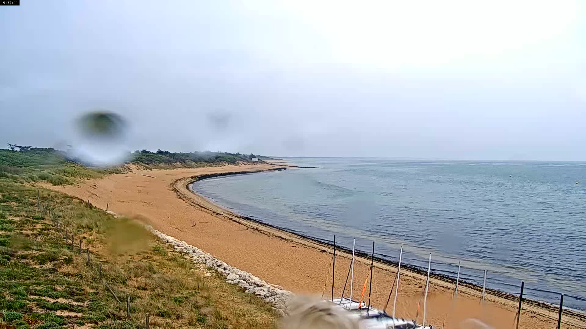 A sandy beach curves along a calm, overcast sea, with a few sailboats beached at the far end.