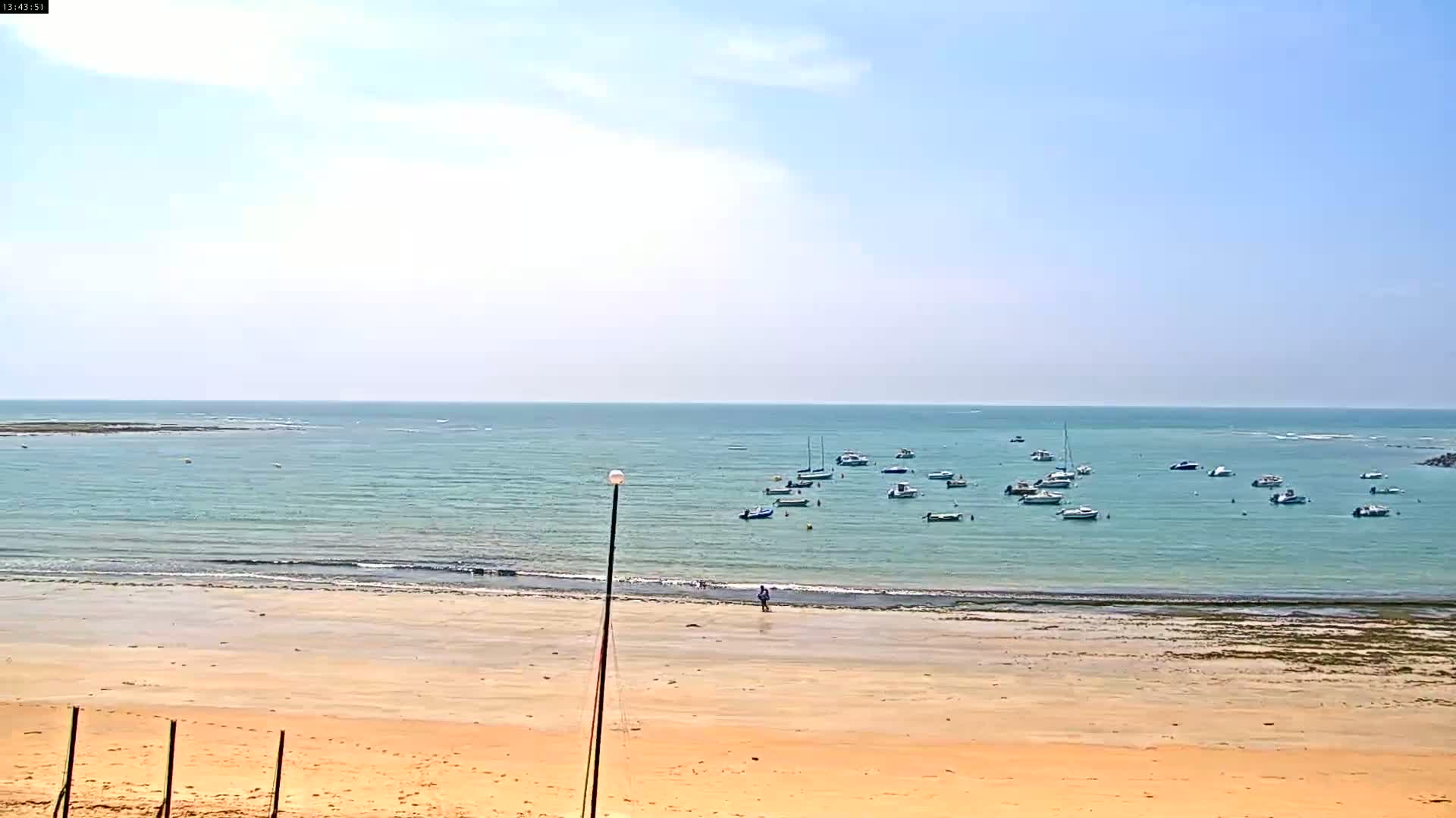 A sandy beach stretches to a calm, light-blue ocean where numerous small boats are anchored under a mostly clear sky.