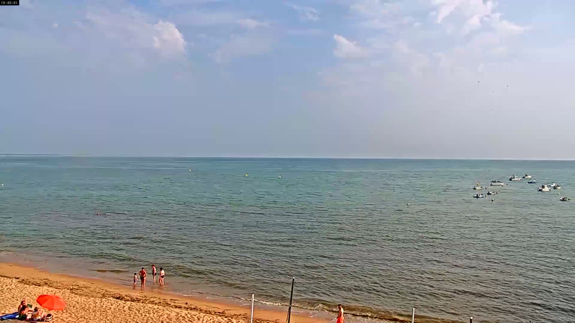 A sandy beach with a few people on it slopes down to a calm sea where several small boats are anchored under a partly cloudy sky.