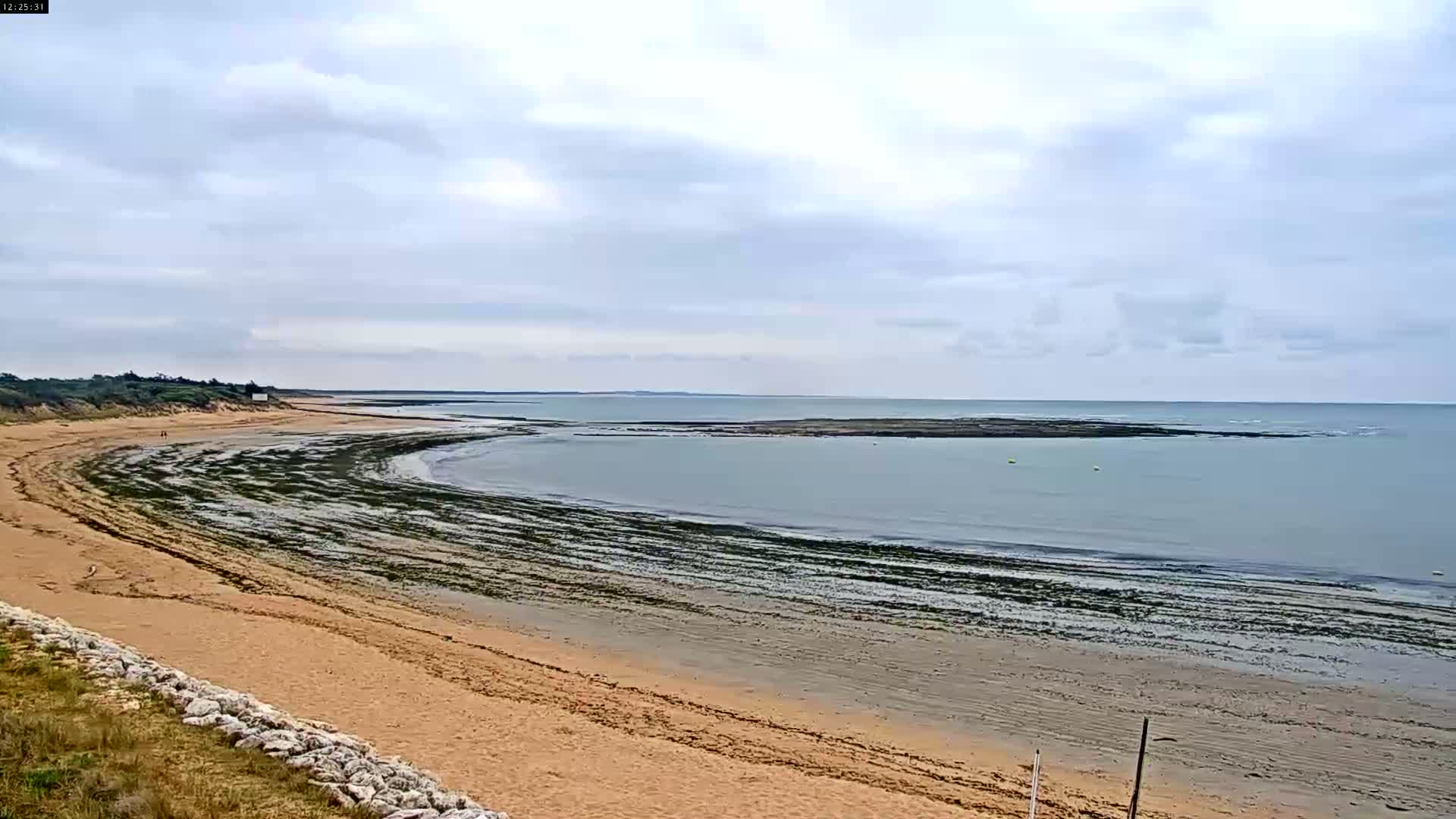 A sandy beach curves around a calm, gray ocean under a cloudy sky.