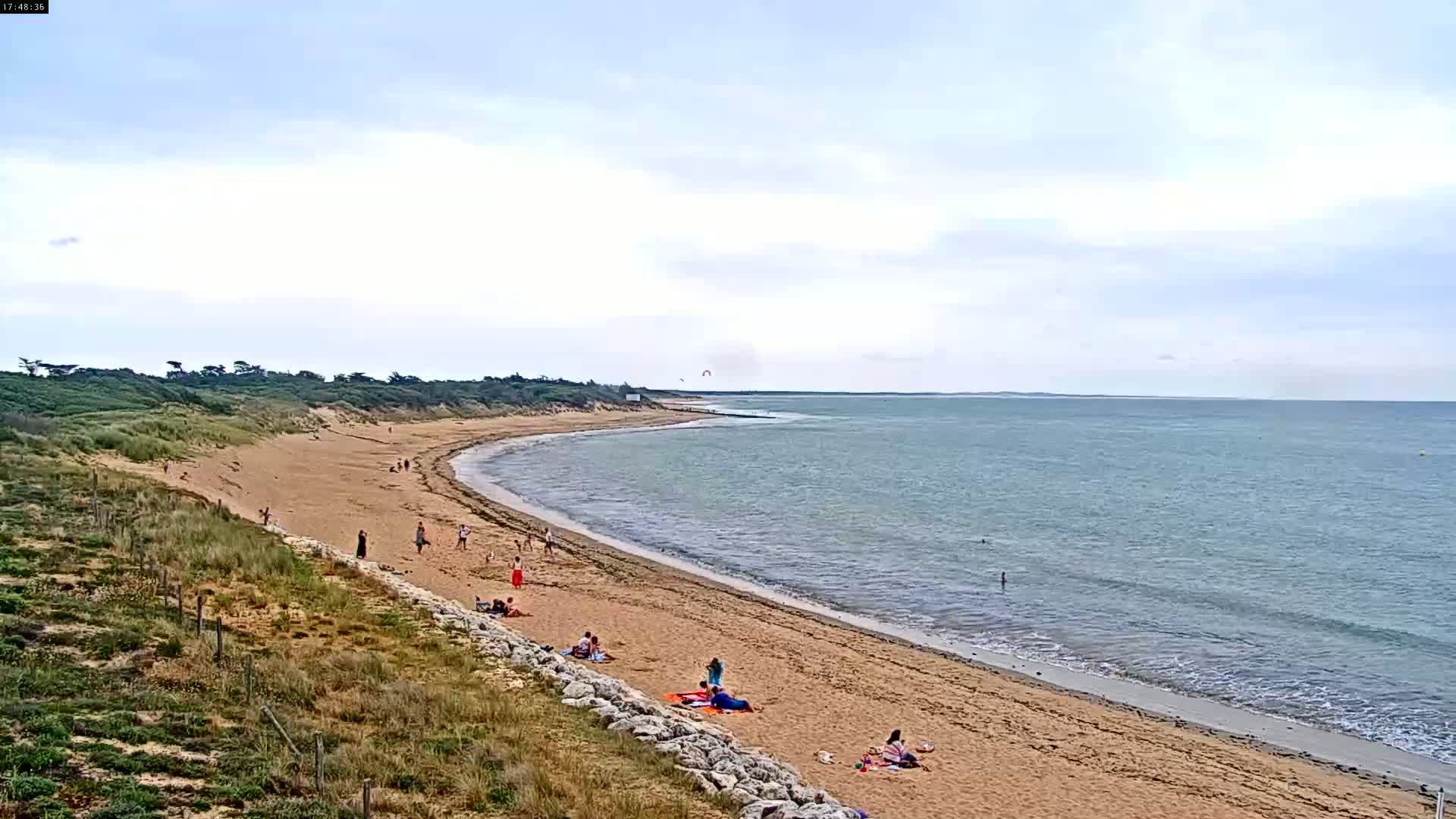A sandy beach curves along a calm ocean under a mostly cloudy sky, with several people scattered along the shore.