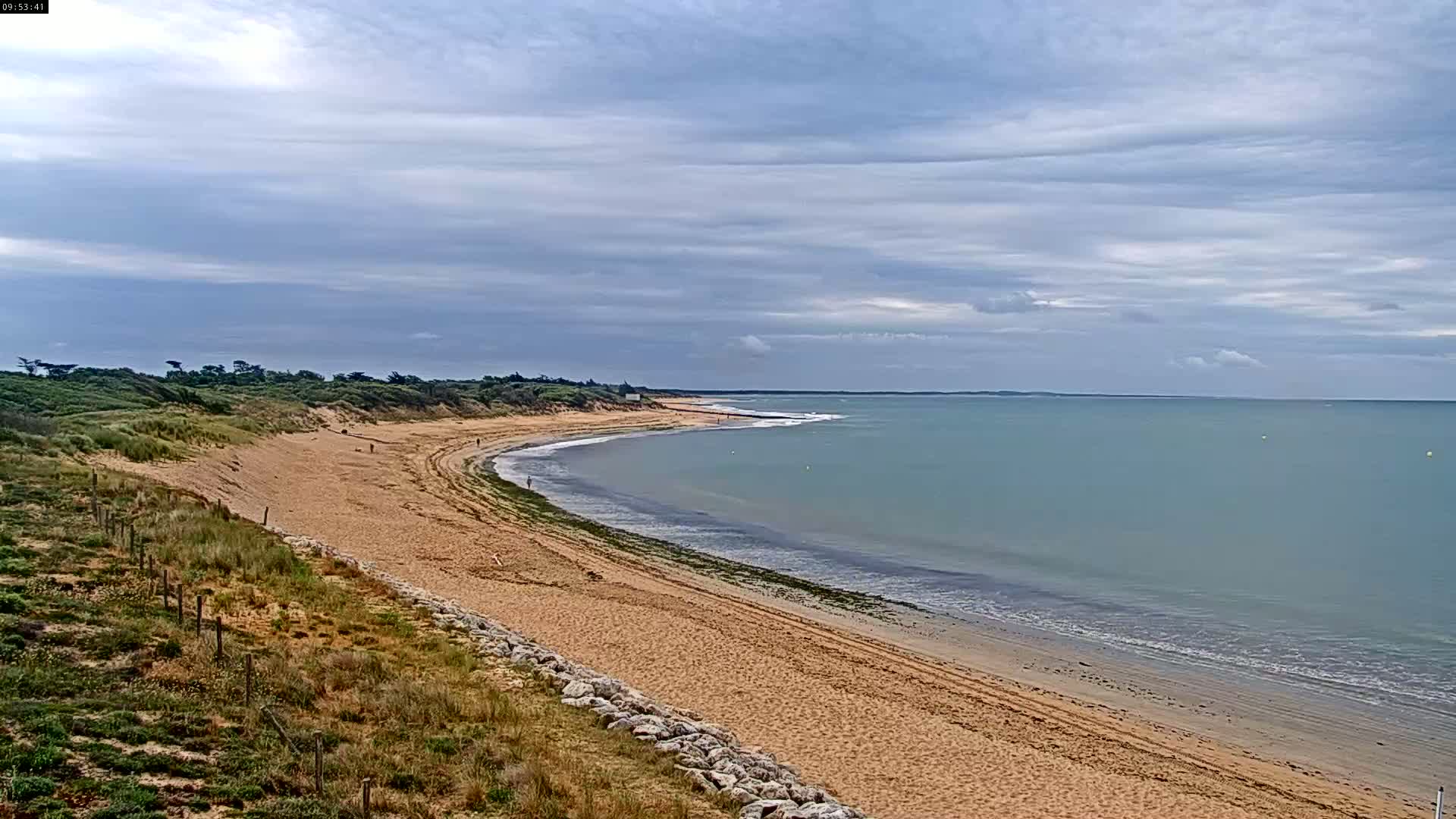 A long sandy beach curves gently around a calm, light-blue ocean under a mostly cloudy sky.