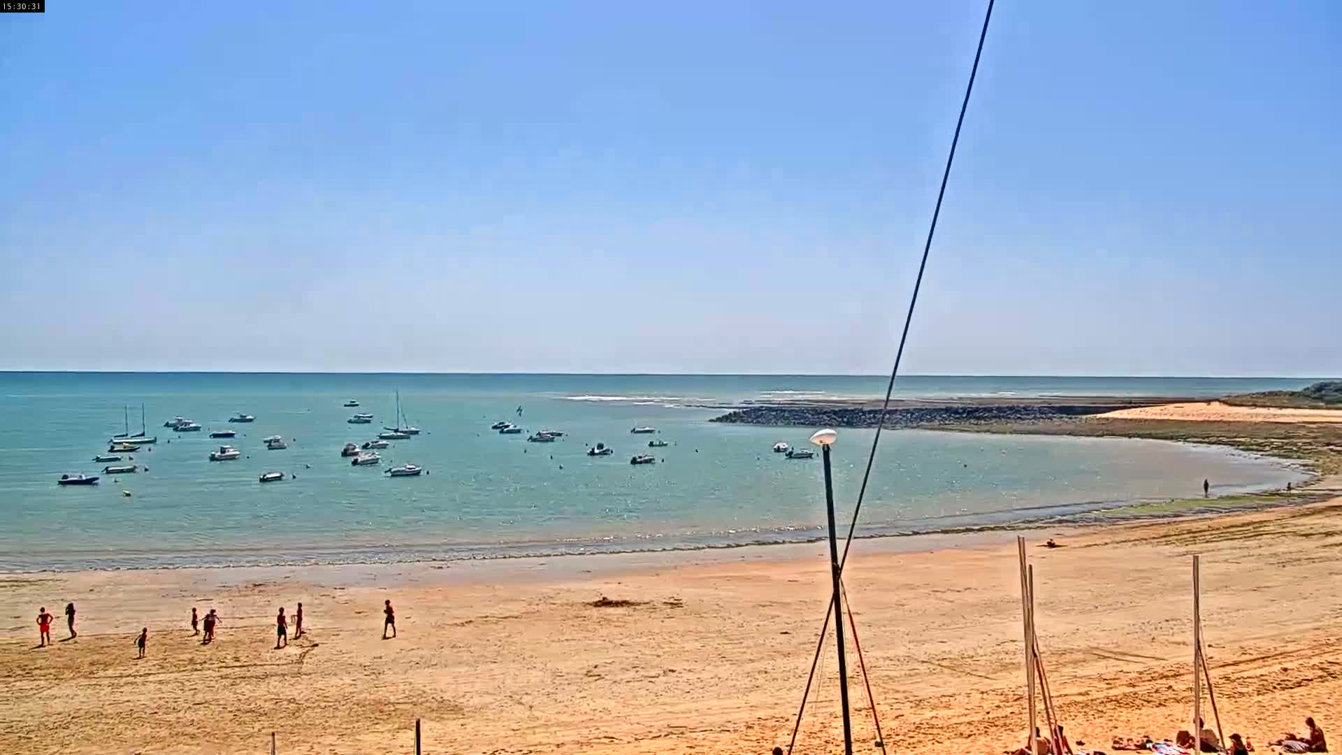 A sandy beach with several people playing on the sand and numerous boats anchored in calm, clear water under a bright, sunny sky.