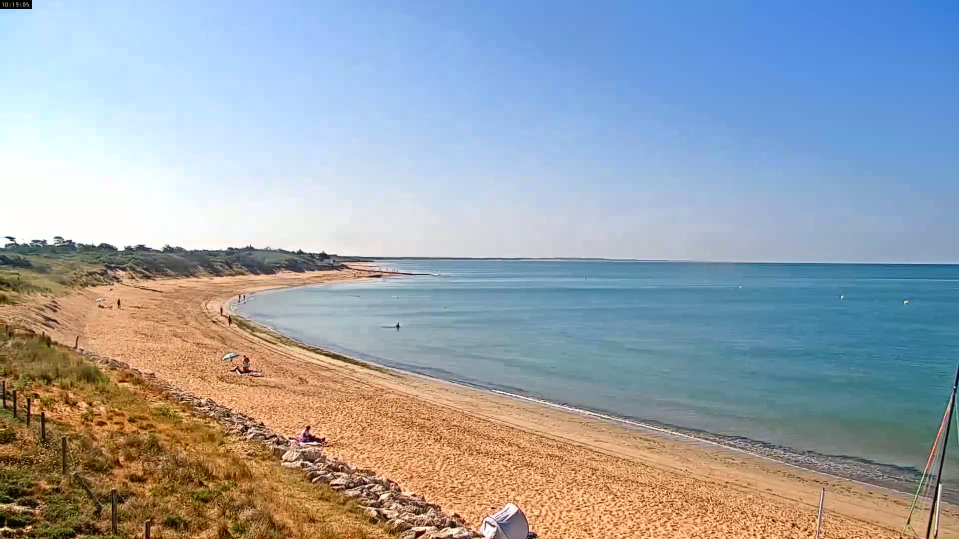 A sandy beach curves along a calm, light-blue ocean under a clear blue sky.