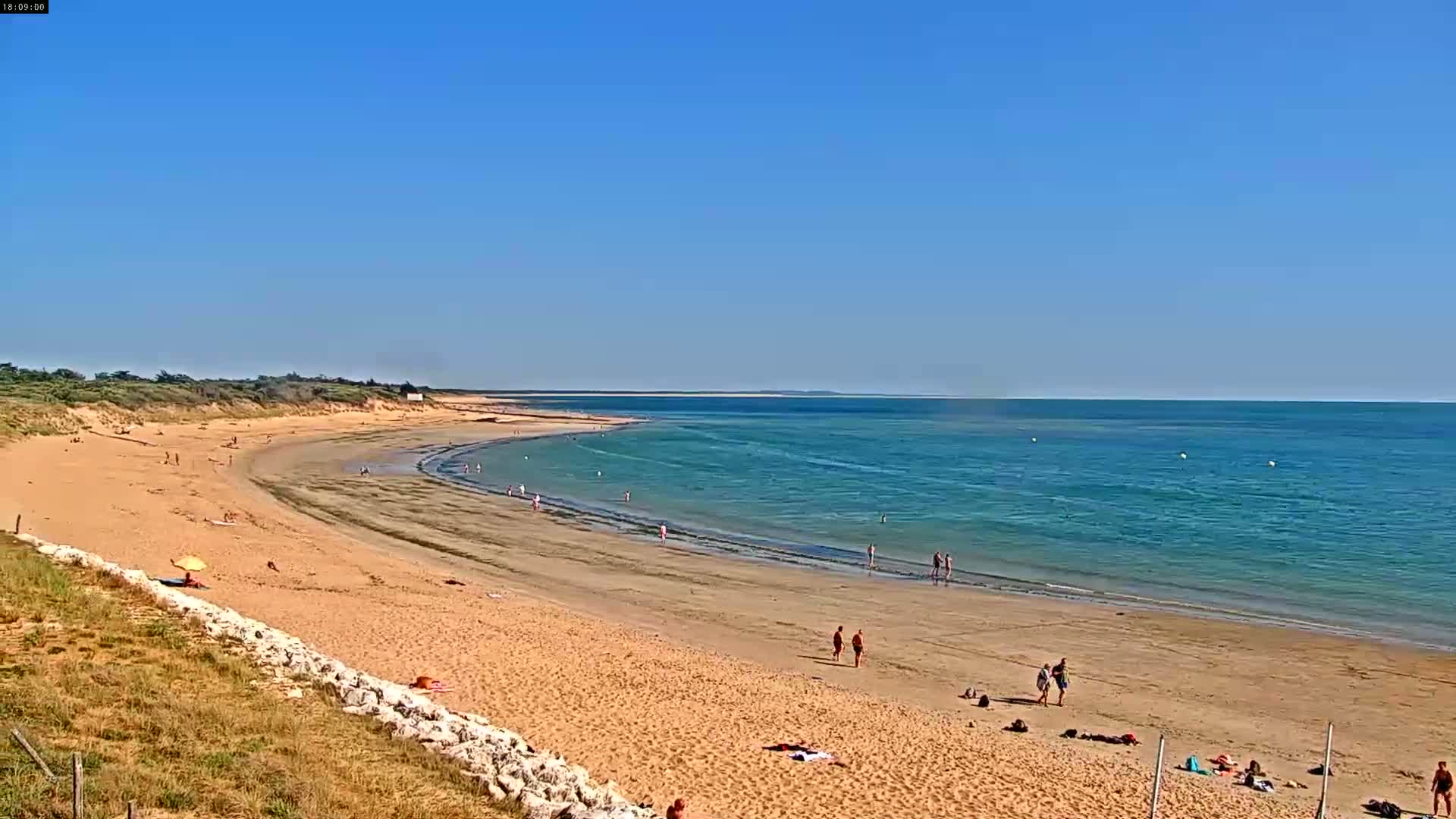 A sandy beach curves gently into a calm, turquoise ocean under a clear blue sky, with a few people scattered along the shore.