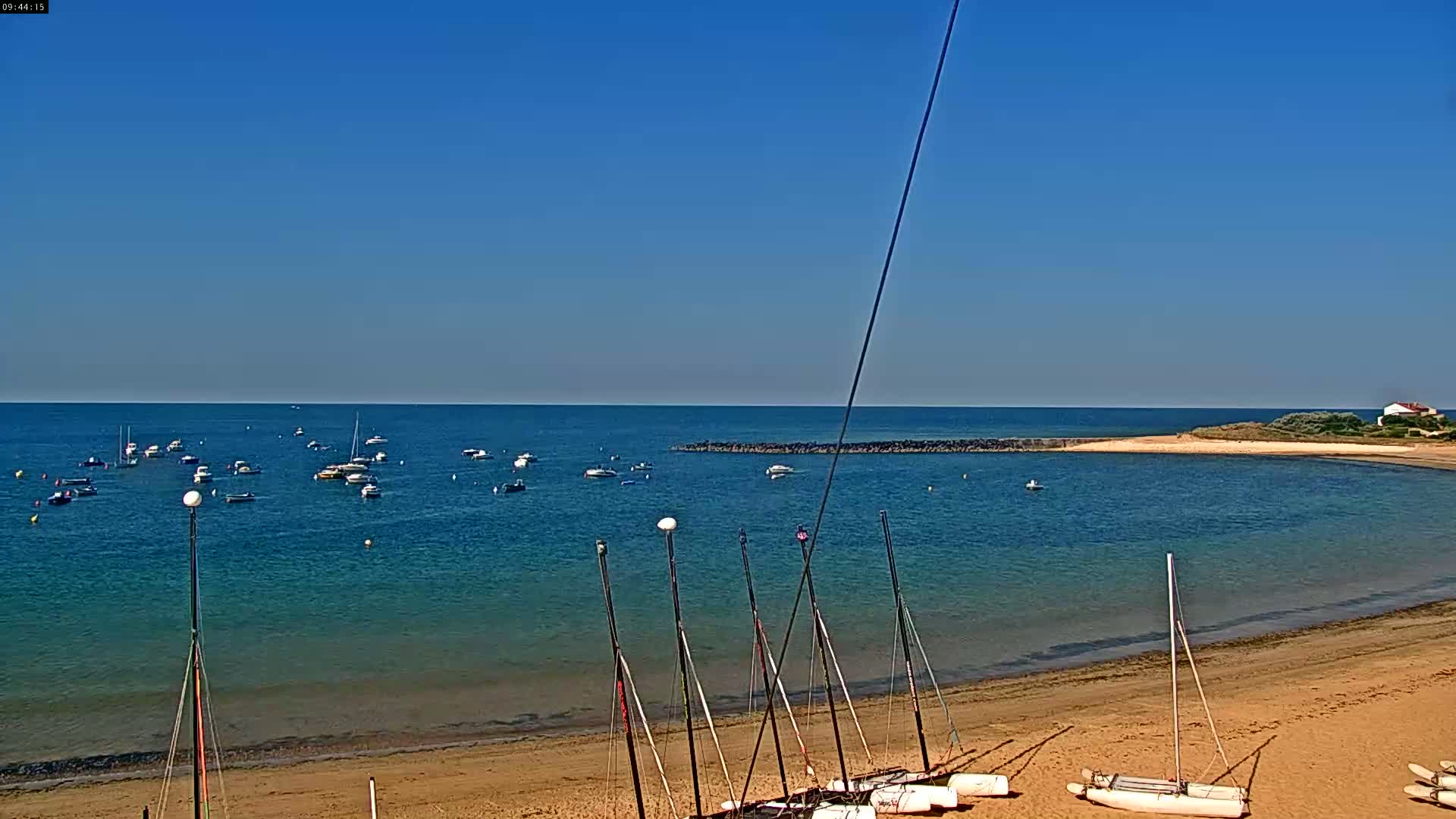 A sandy beach with several small sailboats on the shore and numerous boats in the calm, blue water under a clear, sunny sky.