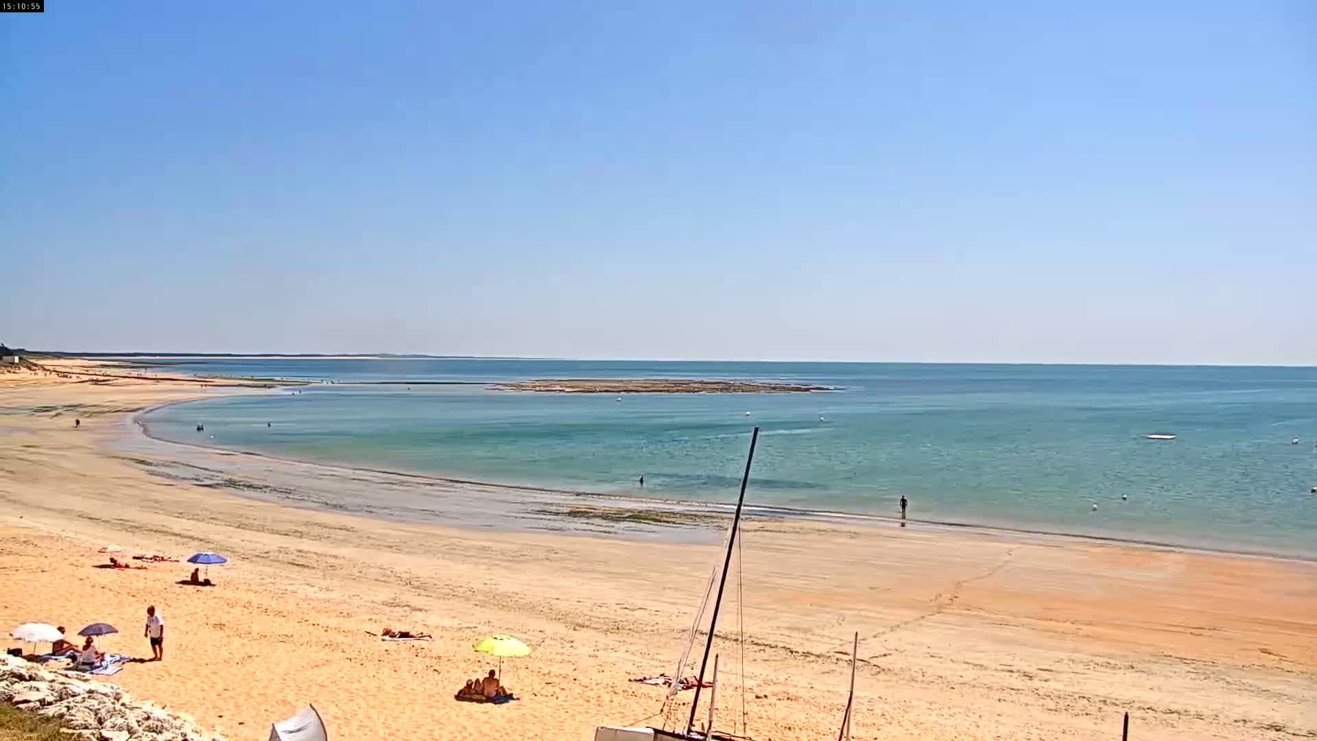 A sandy beach with a few people and umbrellas stretches along a calm, light-green ocean under a clear blue sky.