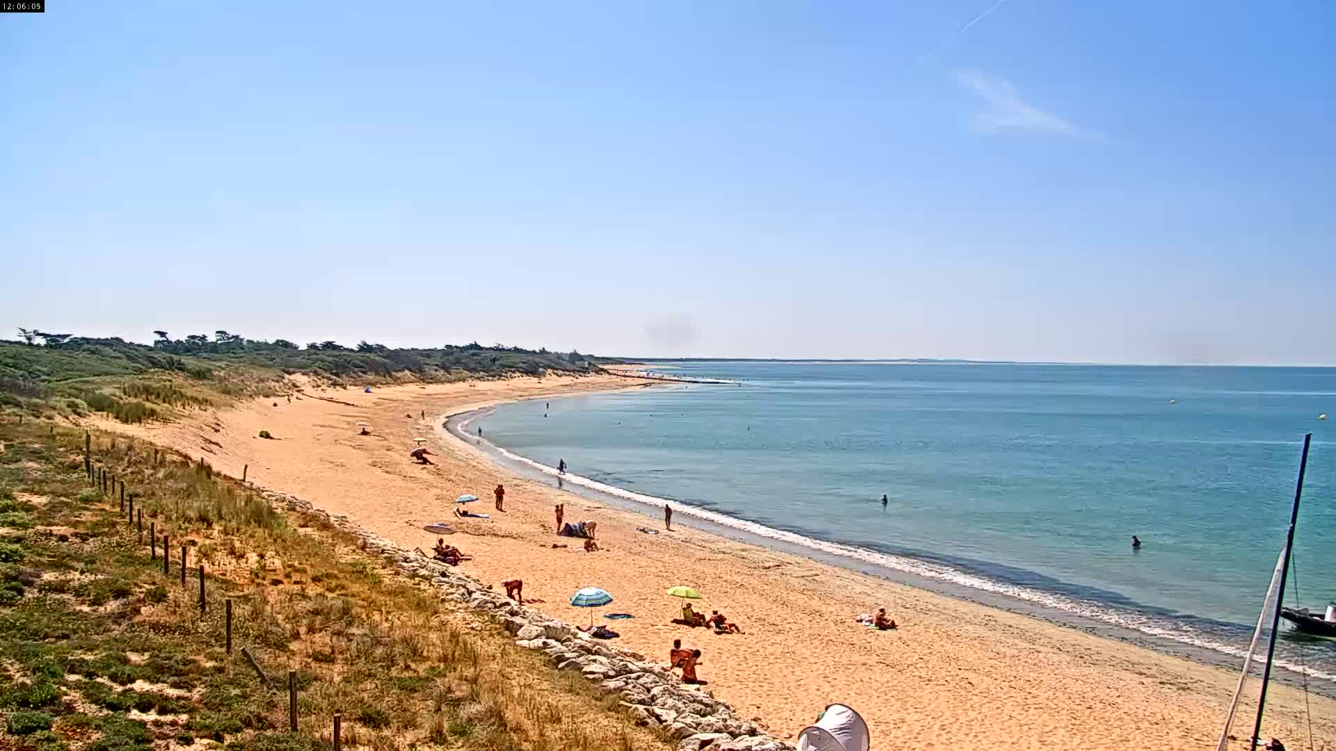A sandy beach curves gently along a calm, light-blue ocean under a clear blue sky, with a few people scattered across the sand and in the water.