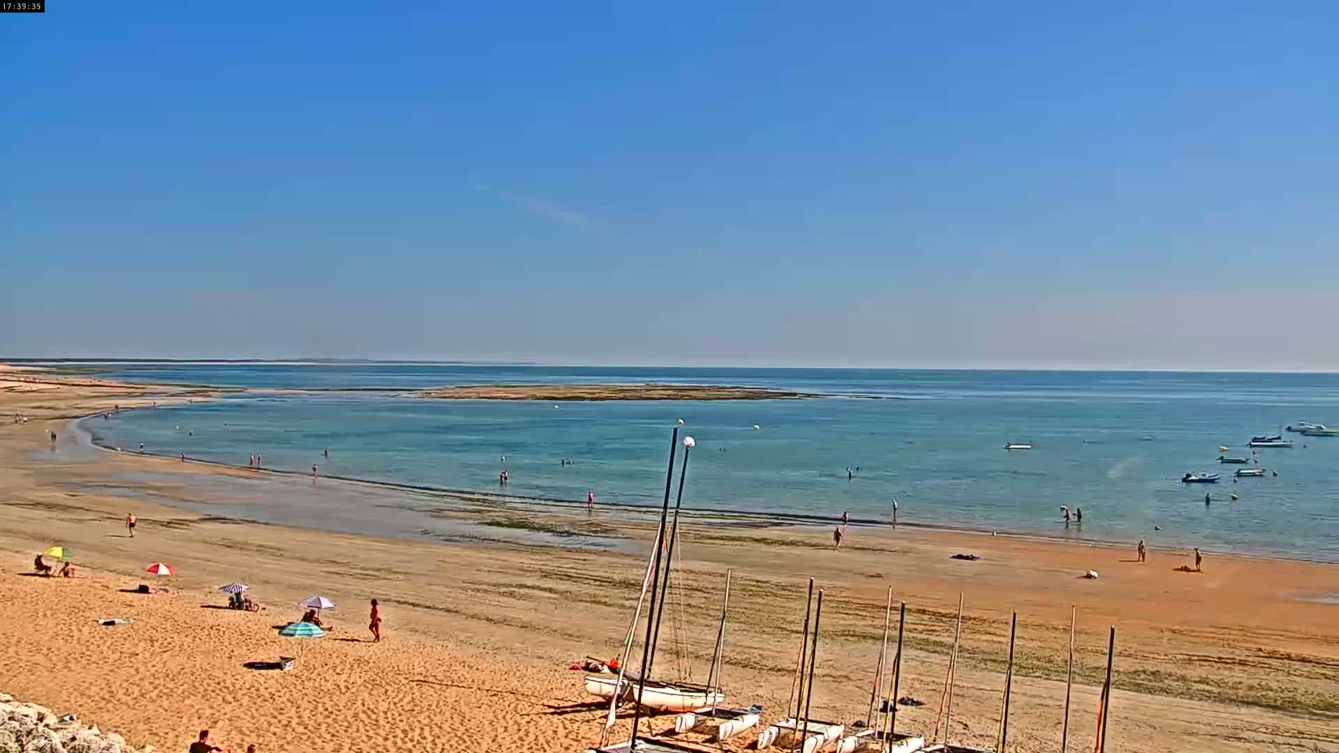 A sandy beach with scattered people and several sailboats rests on a calm, light-blue ocean under a clear blue sky.