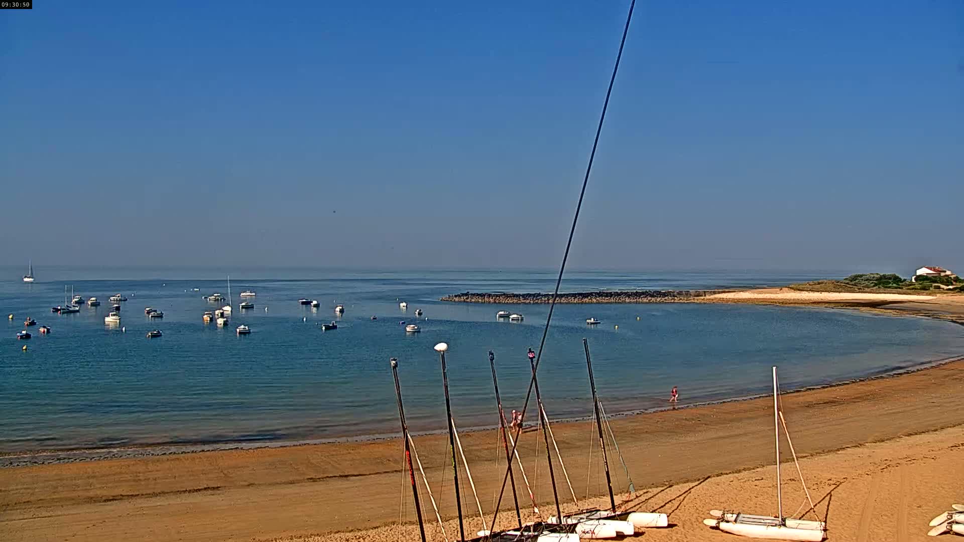 A sandy beach with several sailboats on the shore and numerous small boats anchored in calm, clear water under a clear blue sky.