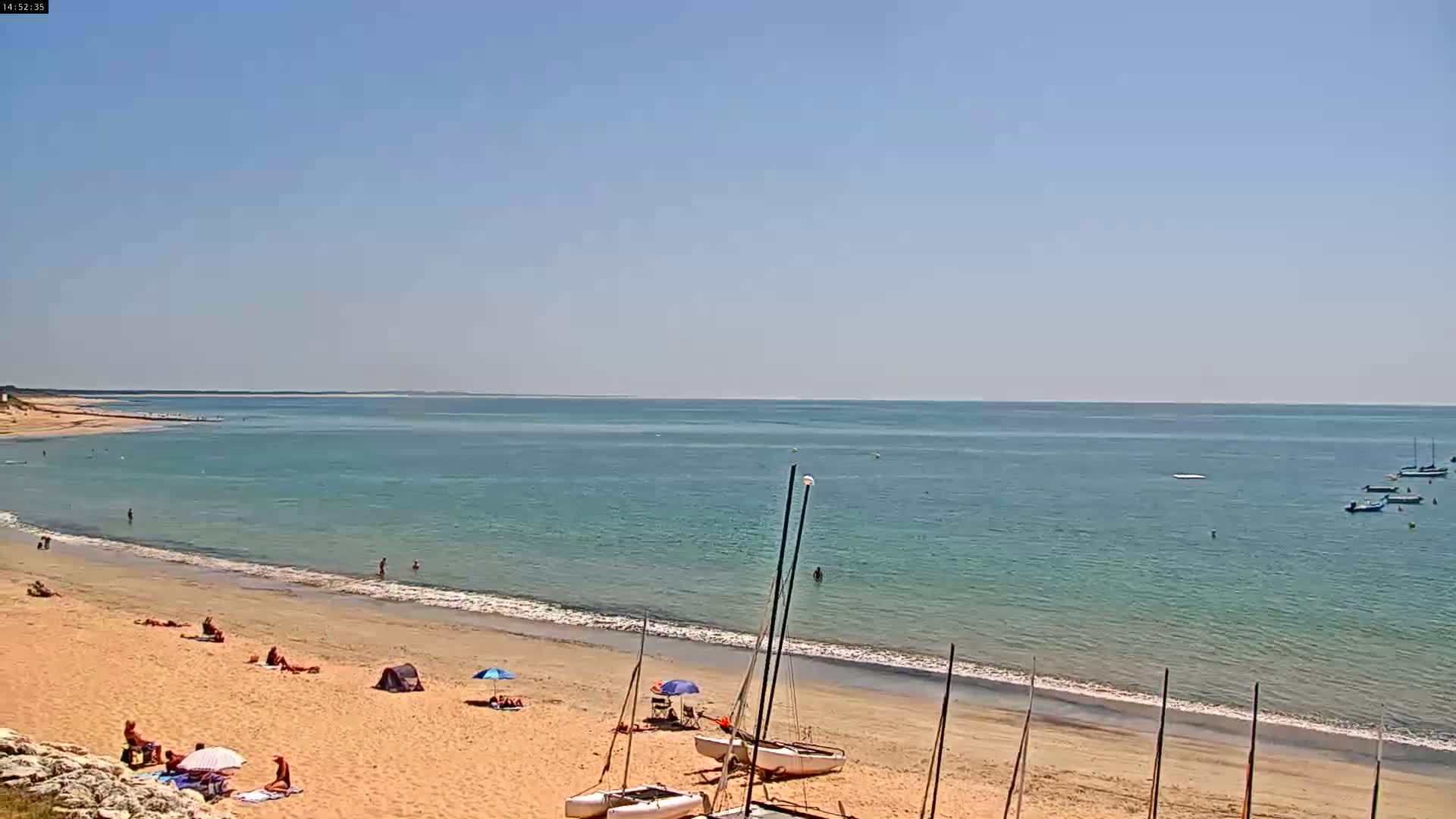 A sandy beach with several people sunbathing and swimming, boats in the calm, clear water, and sailboats on the shore under a clear blue sky.