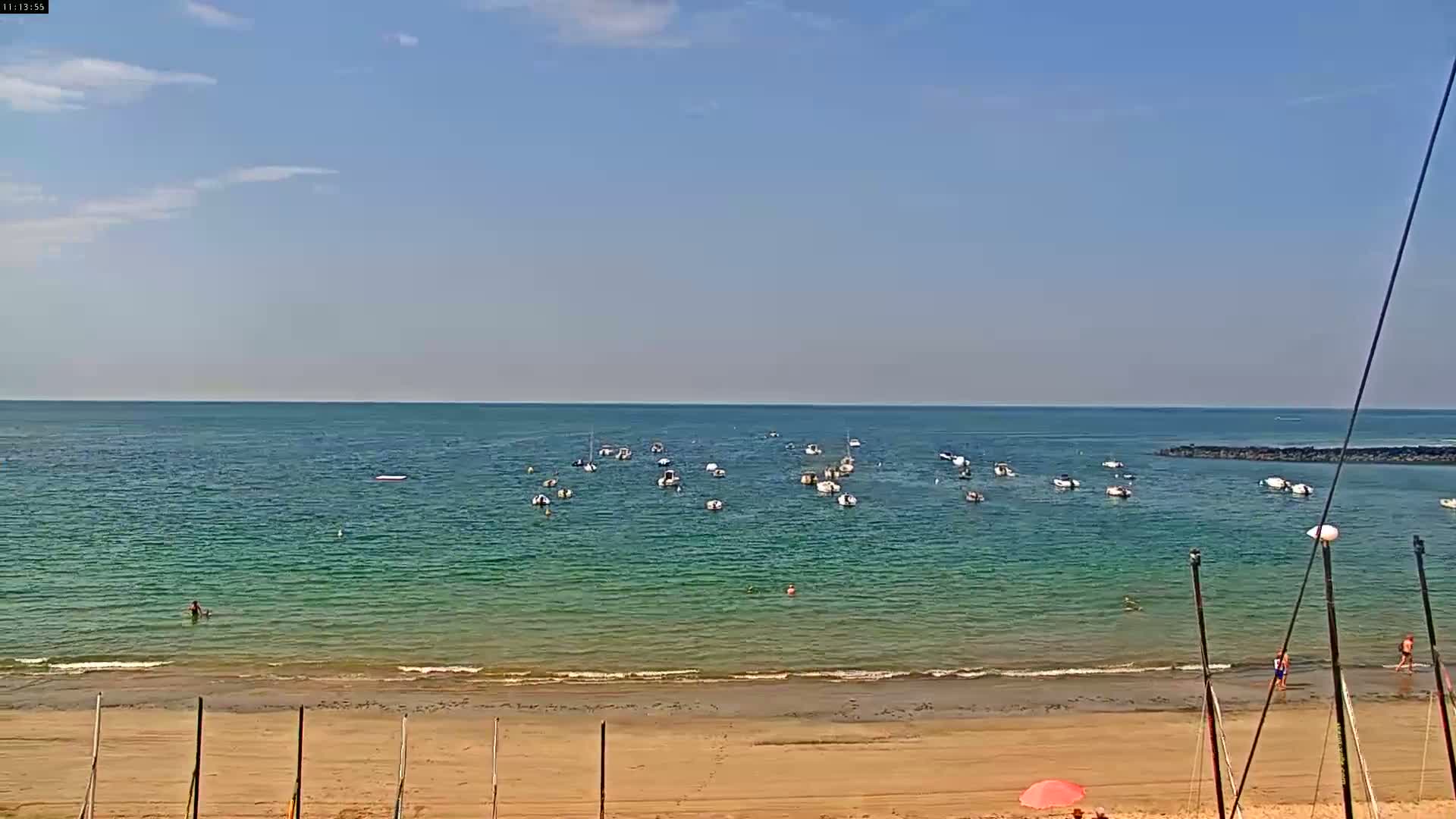 A sandy beach with calm, clear teal water and numerous small boats anchored offshore under a mostly sunny sky.