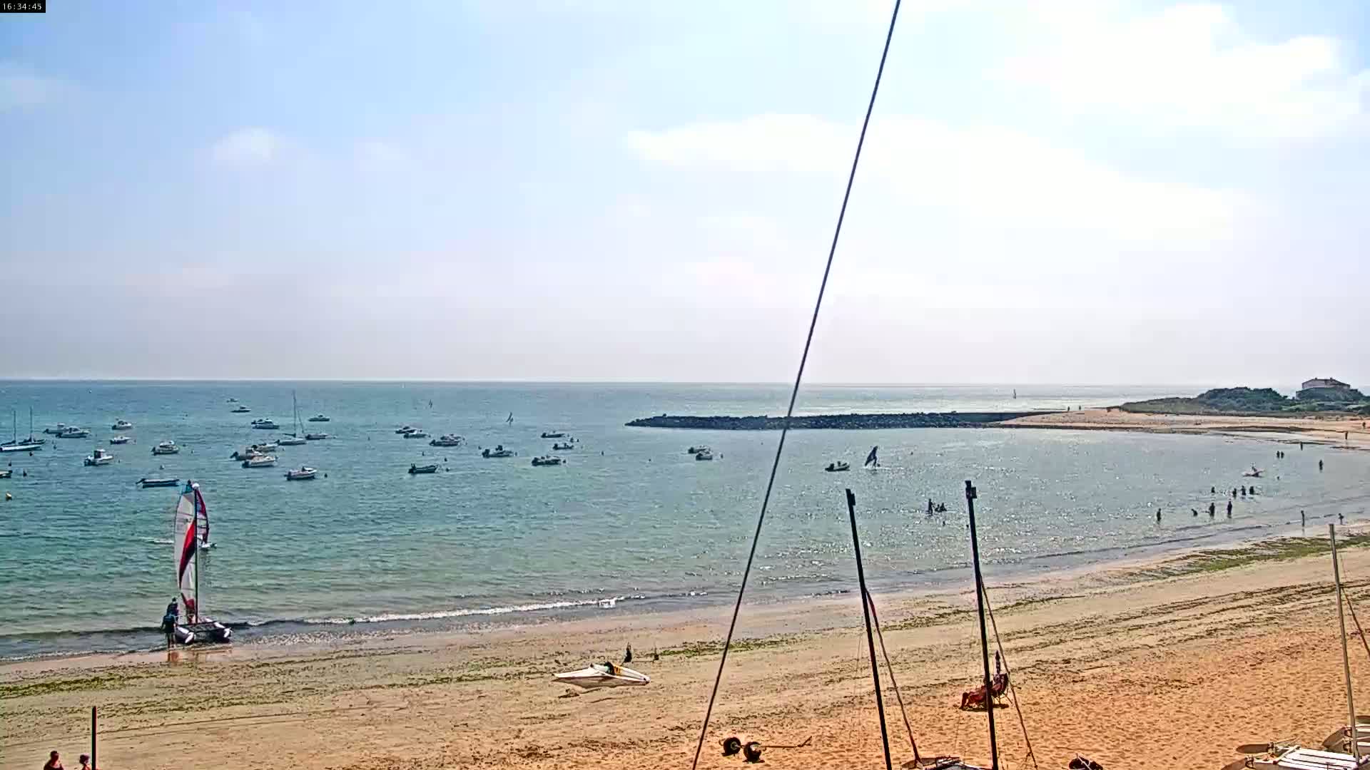 A sandy beach with calm, light-blue water and numerous small boats and people swimming and on the sand under a mostly clear, bright sky.