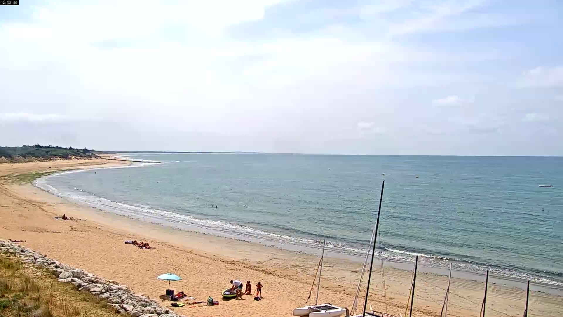 A sandy beach with calm ocean waters under a partly cloudy sky, showing several people relaxing and swimming.