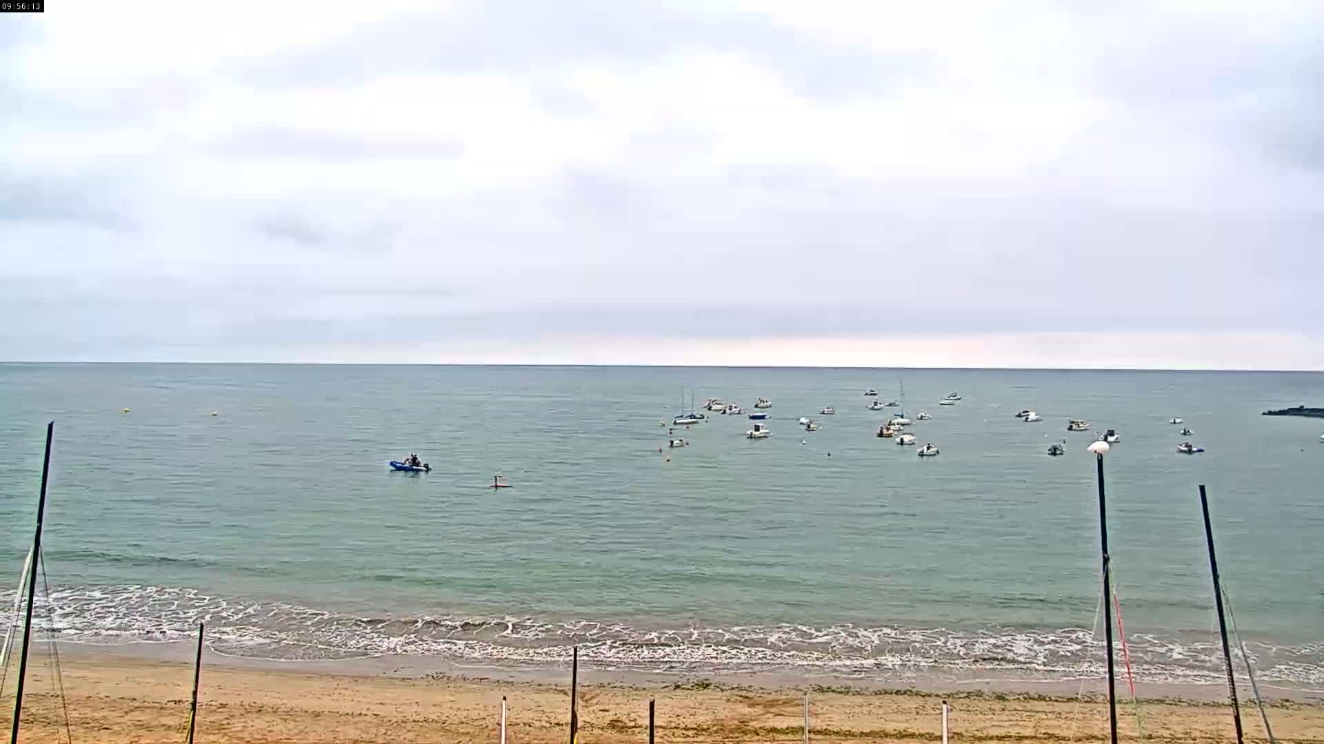 A sandy beach with calm, light-green water, numerous small boats, and an overcast sky.