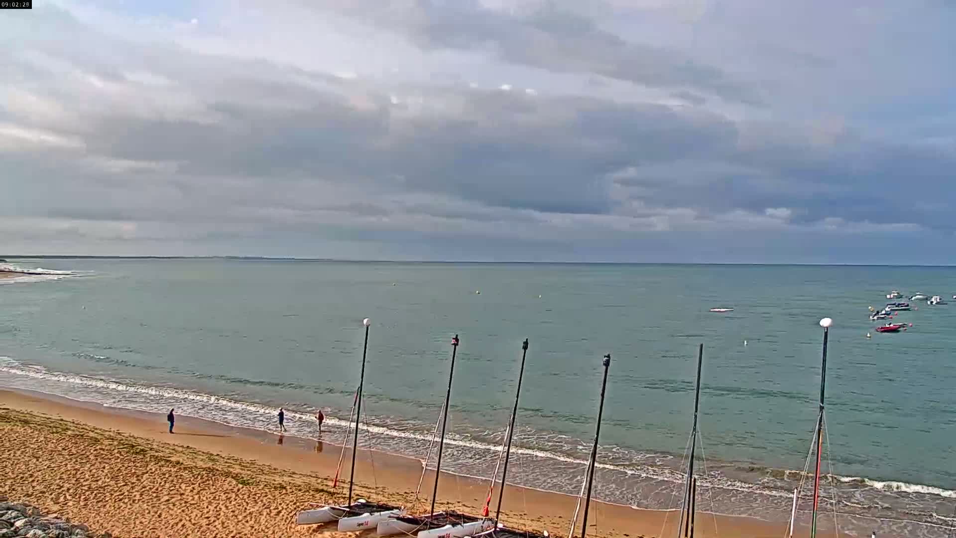 A sandy beach with several small sailboats on the shore and a few people walking along the waterline under a mostly cloudy sky.